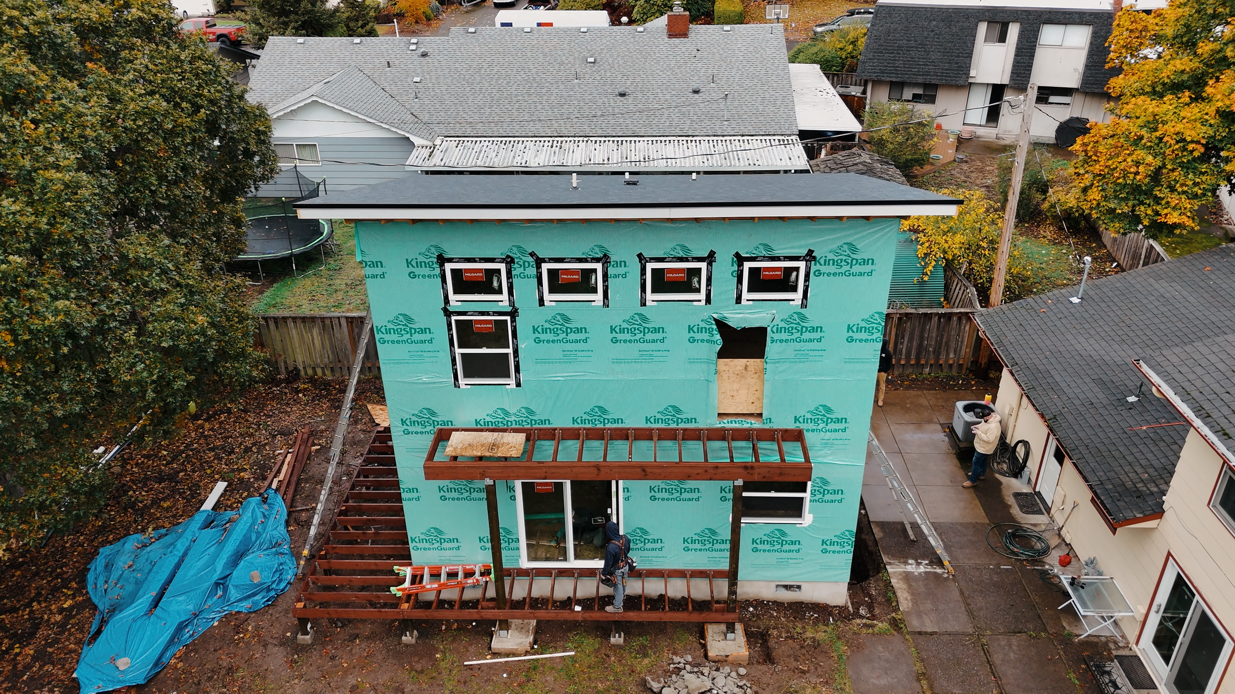 Two-story house under construction with Kingspan GreenGuard insulation, wooden framework for a porch or awning, and workers on site.