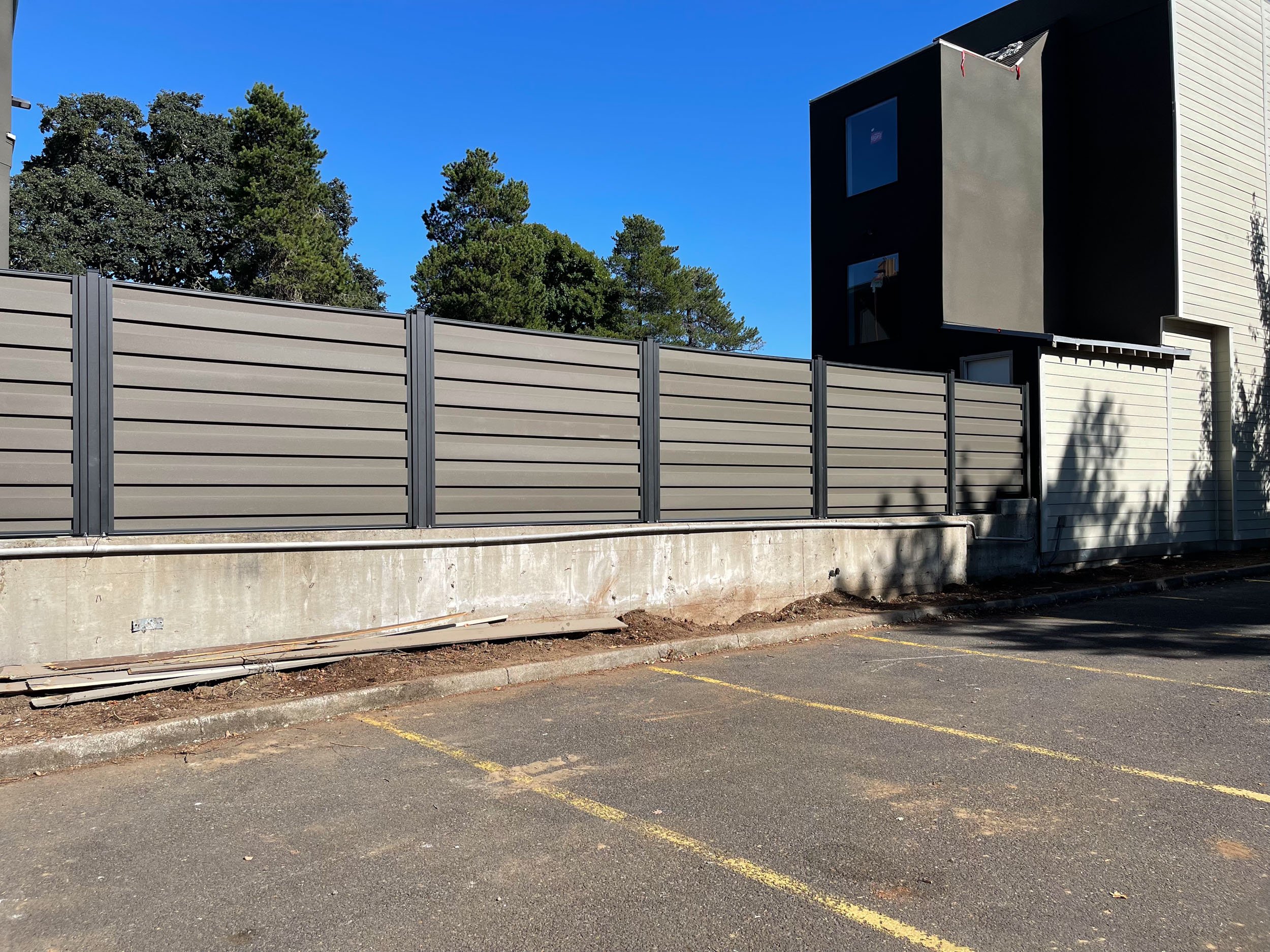 A modern building with a gray horizontal panel fence in the foreground and a paved area with yellow parking lines. Trees are visible in the background under a clear blue sky.