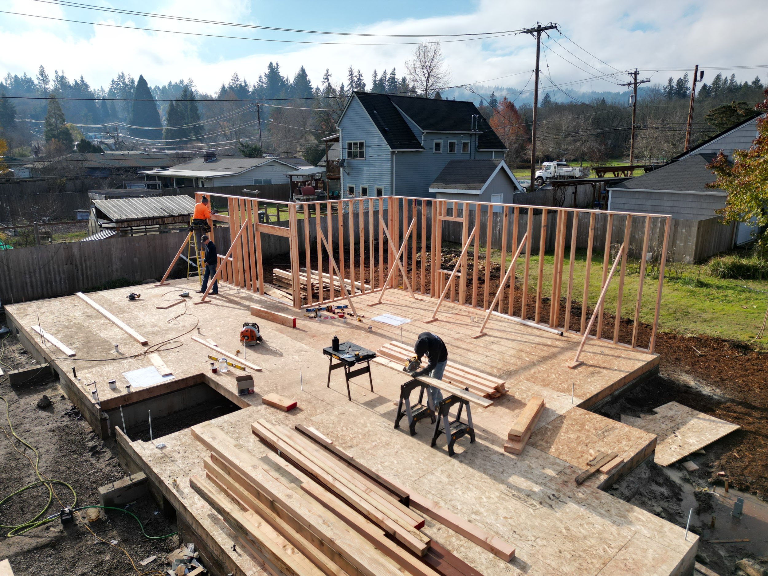 Two construction workers building a wooden house frame on a residential property, with tools and lumber scattered on the plywood base. A partially constructed wall is visible with wooden studs, surrounded by neighboring houses and trees.