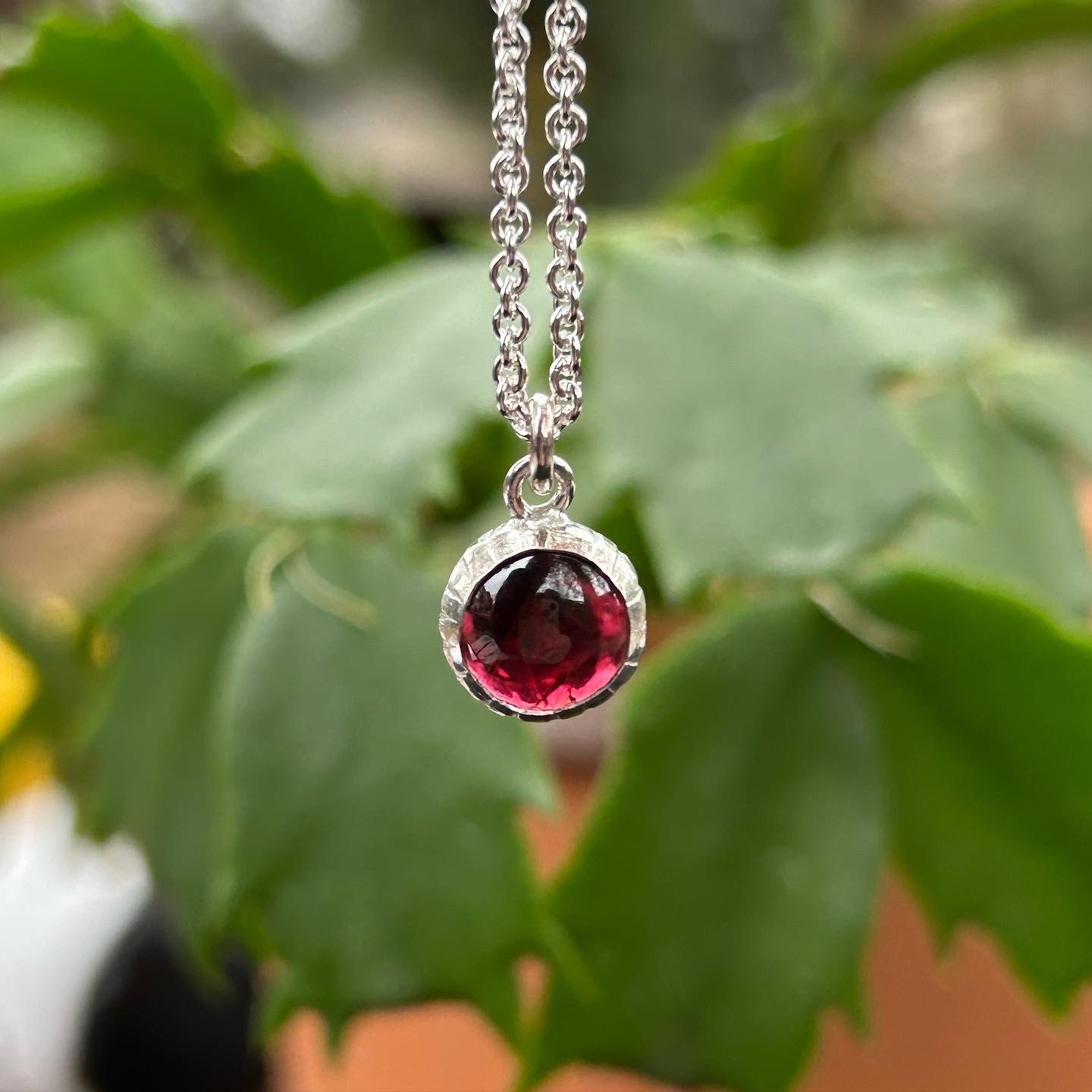 A circular garnet cabochon is set in a silver necklace, dangling in front of a blurred plant