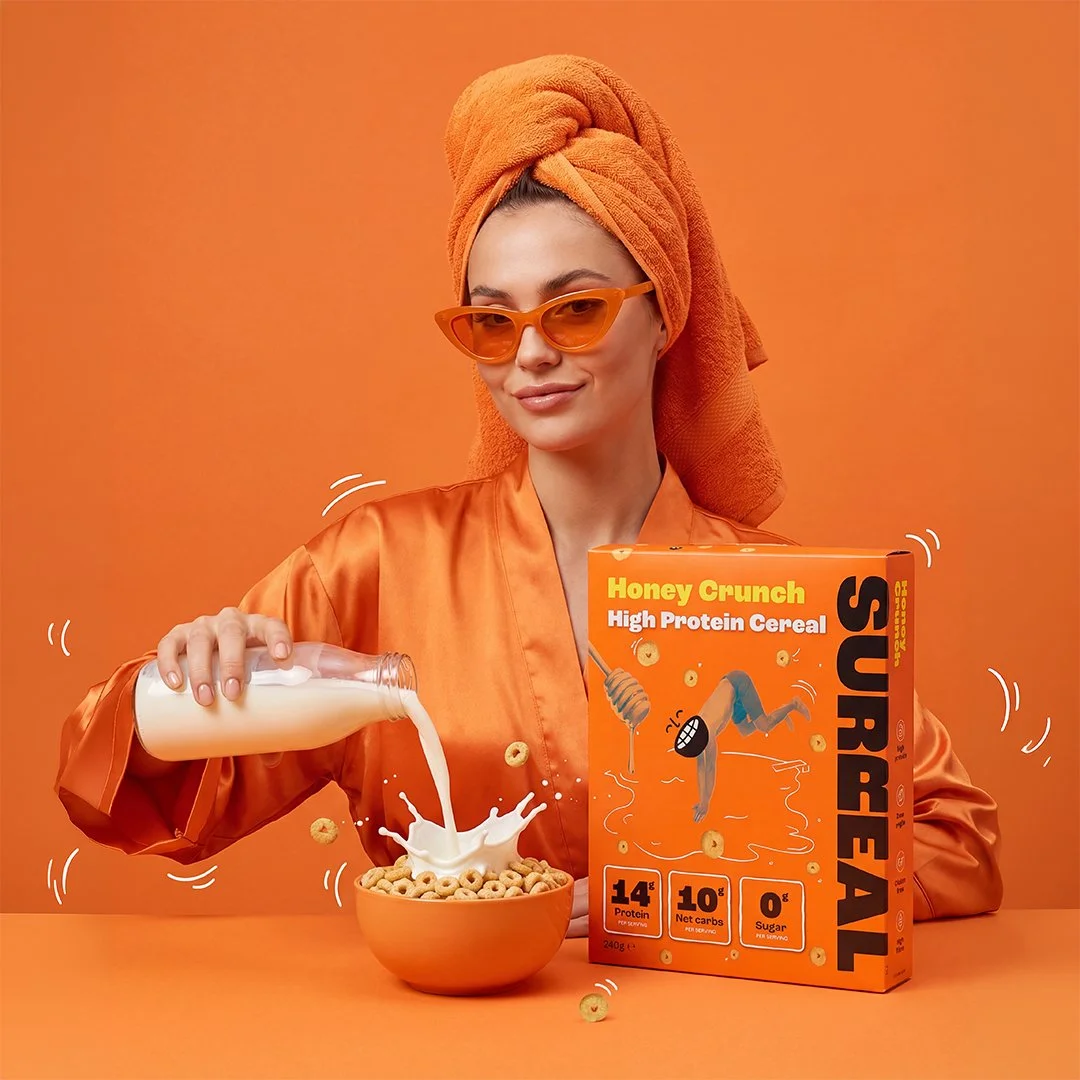 Lifestyle product image of Honey Crunch high protein cereal with milk being poured into a bowl beside the cereal box on a monochrome orange set.