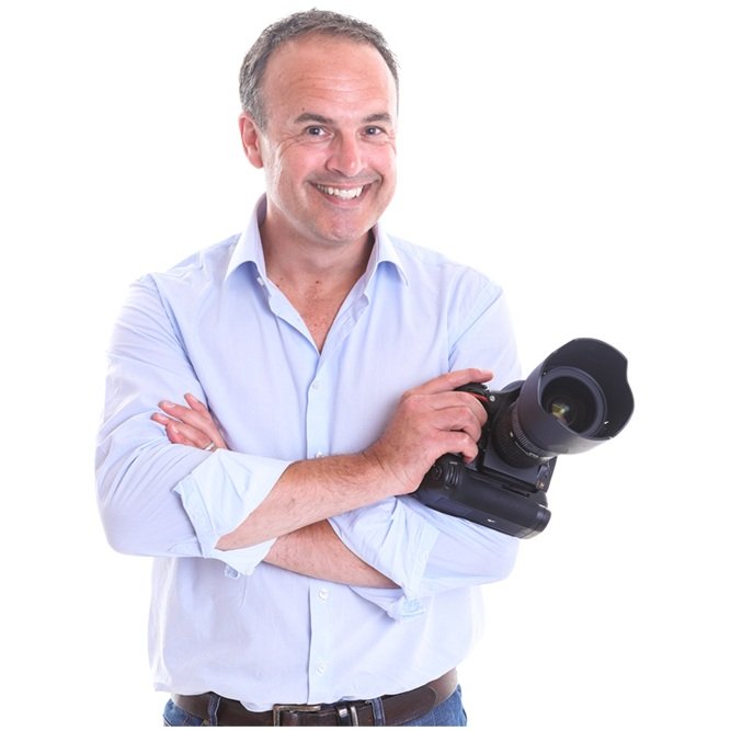 Smiling man in a light blue shirt holding a professional camera with a large lens.