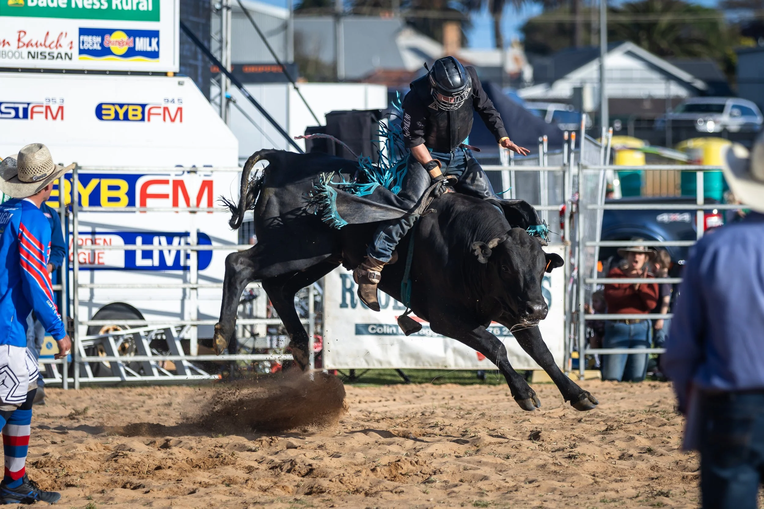 A cowboy riding a bucking black bull during a rodeo event on a dirt arena, with spectators and banners in the background.
