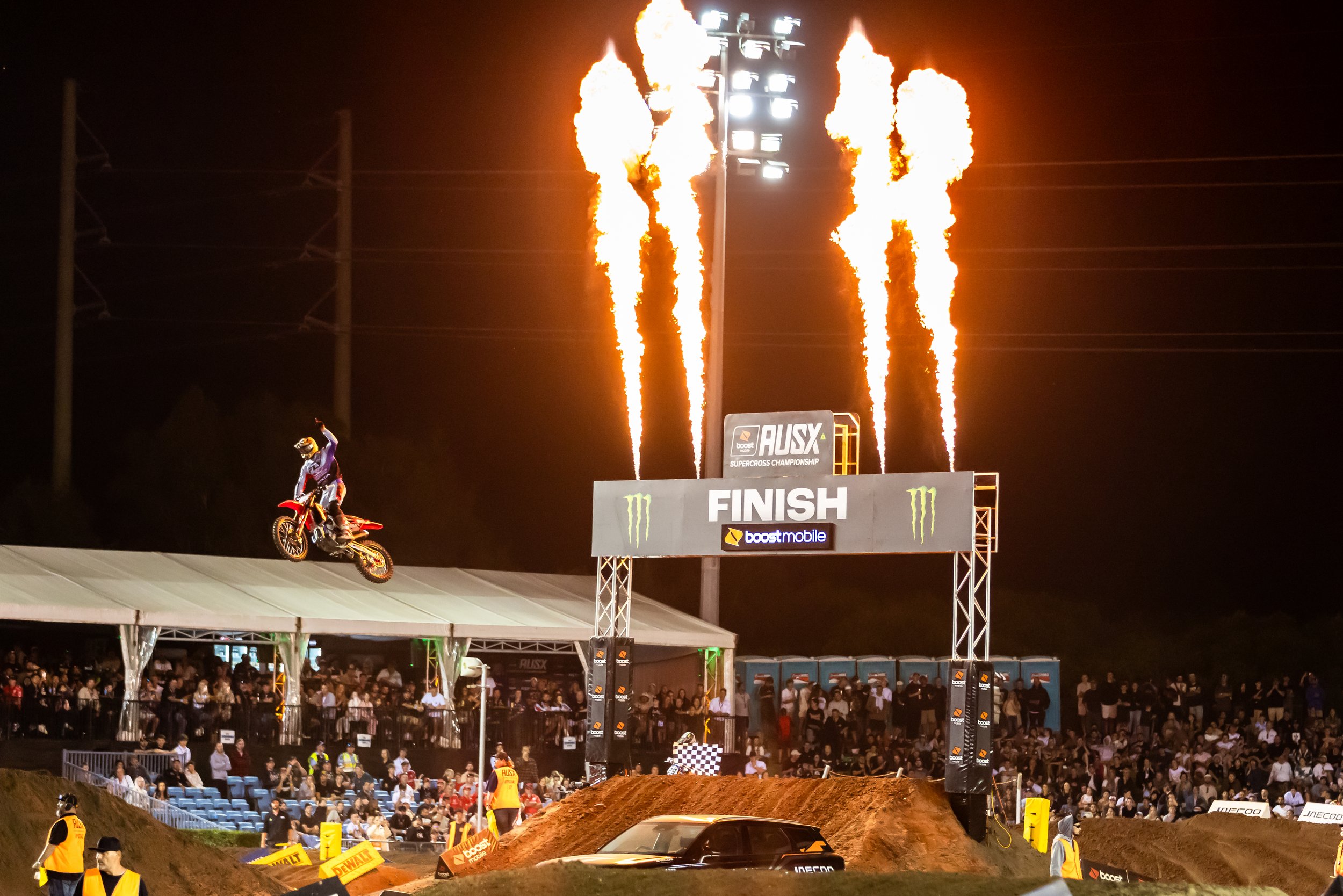 Motocross rider performing a jump at night during a race, with flames shooting from the lighting tower above, a large crowd watching, and a finish banner in the background.