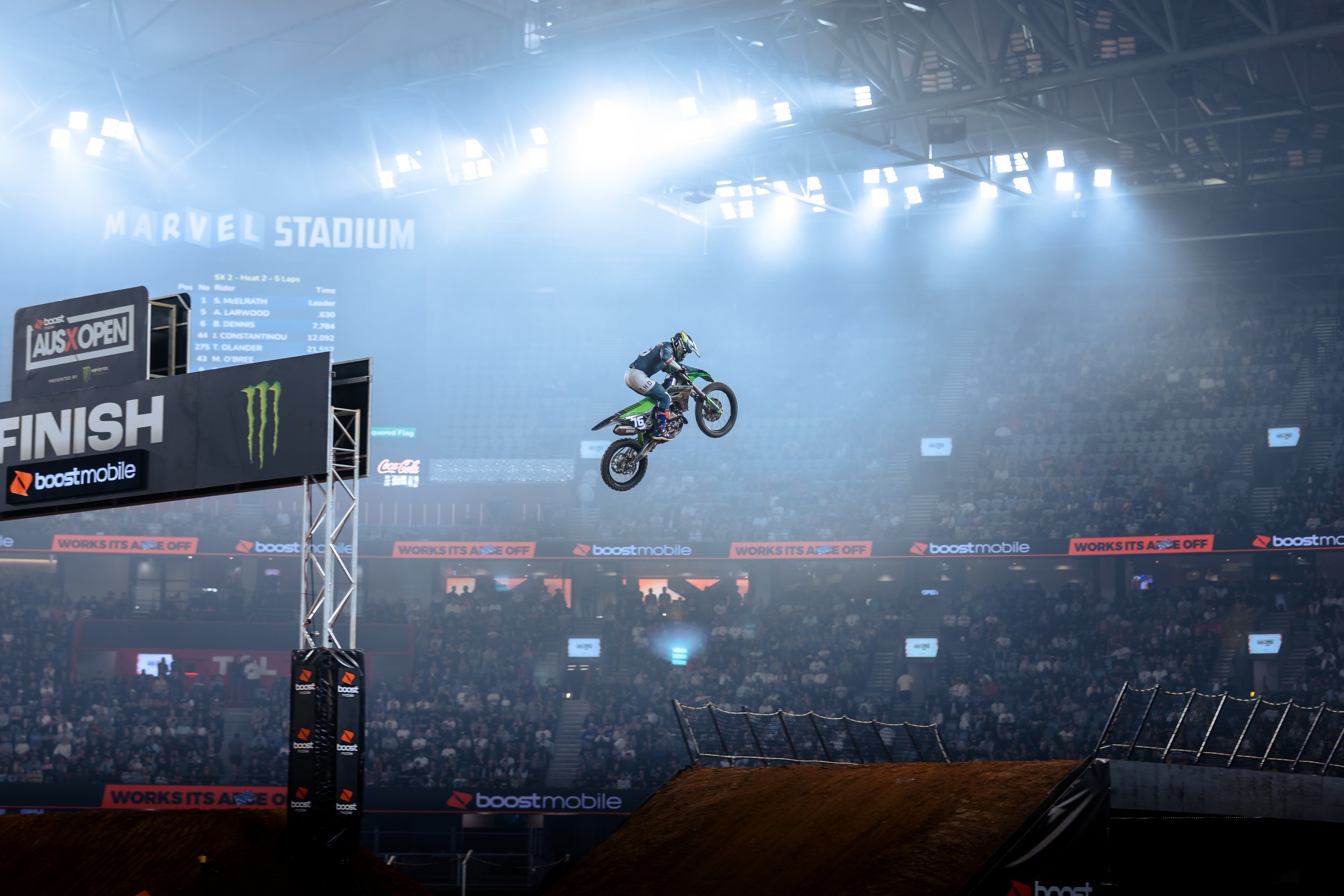 Motocross rider mid-air during a stunt or race inside an indoor stadium with bright lighting and a large audience in the background.