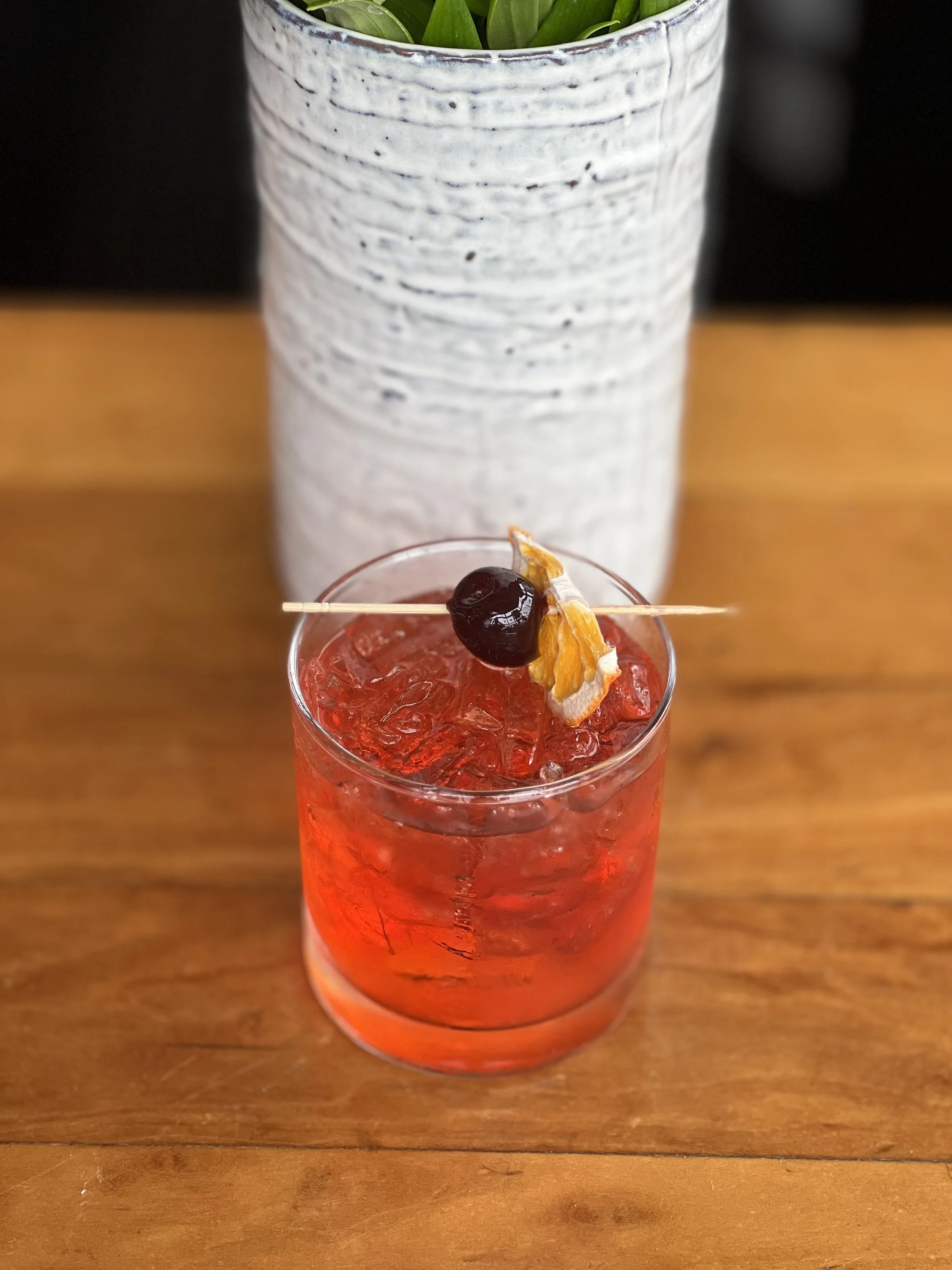 A red cocktail drink with ice, garnished with a cherry and dried fruit slice, in a glass on a wooden table, with a white textured ceramic vase with green plant leaves in the background.