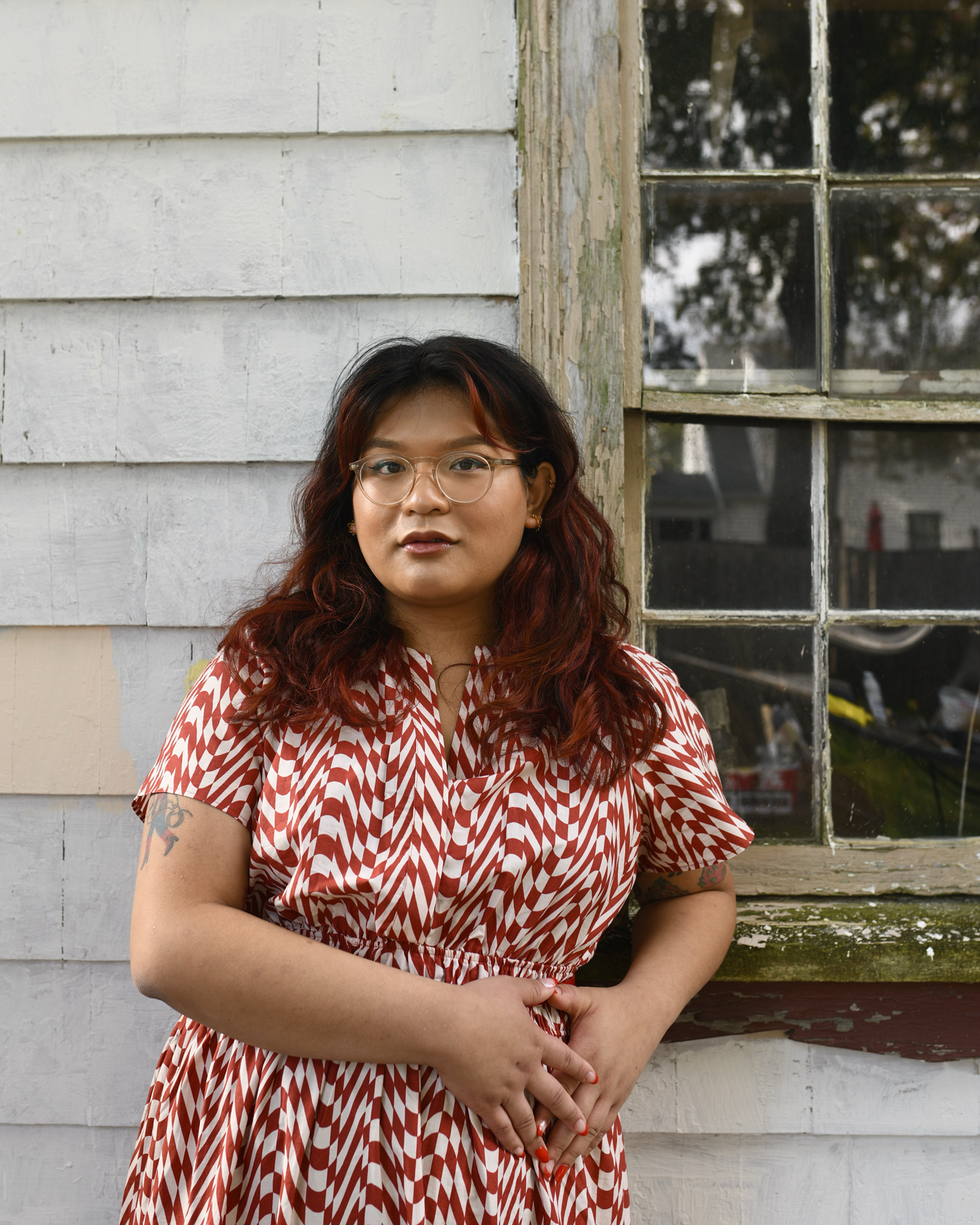 The poet Chrysanthemum stands outdoors and faces the camera with a wall and window behind her. She has golden skin and chest-length dark hair, and wears translucent glasses with a red-and-white patterned dress. Photo by Zach Oren.