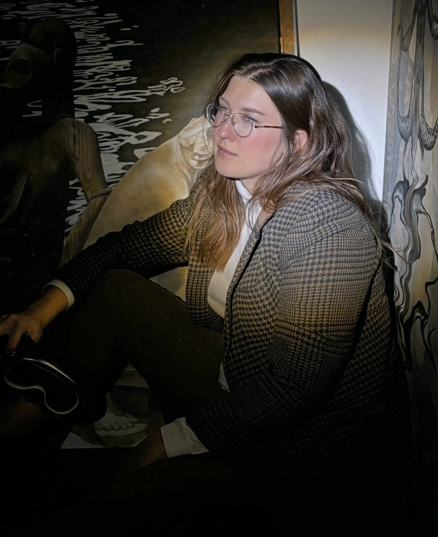 A woman with long brown hair, wearing glasses, a houndstooth blazer, white shirt, and black pants, sitting indoors next to a wall with artwork and antiquities, looks to the left.