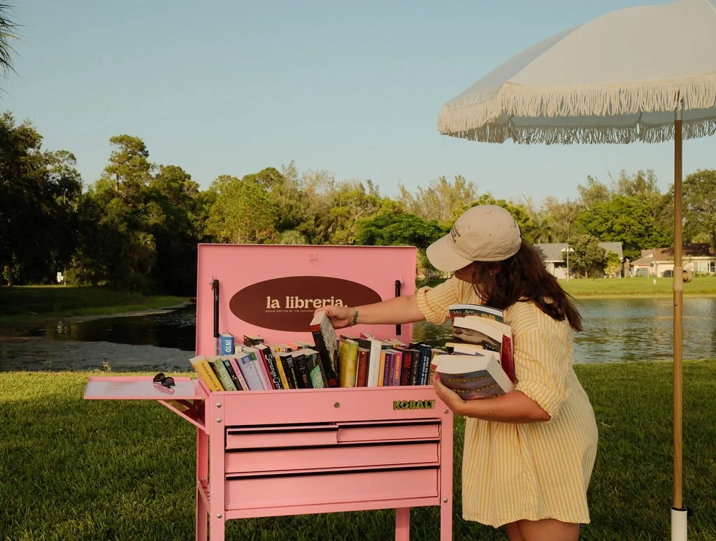 A woman in a yellow striped dress and beige cap browsing books from a pink outdoor book exchange station near a lake with houses and trees in the background, shaded by a cream-colored umbrella.