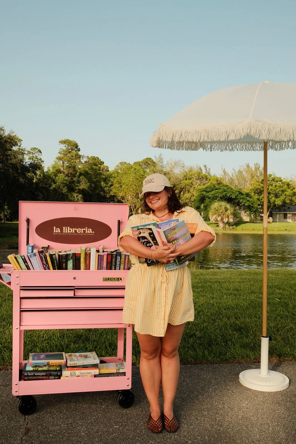 A woman standing outdoors next to a pink mobile library cart filled with books, holding several books and smiling. There is a white patio umbrella and trees in the background.