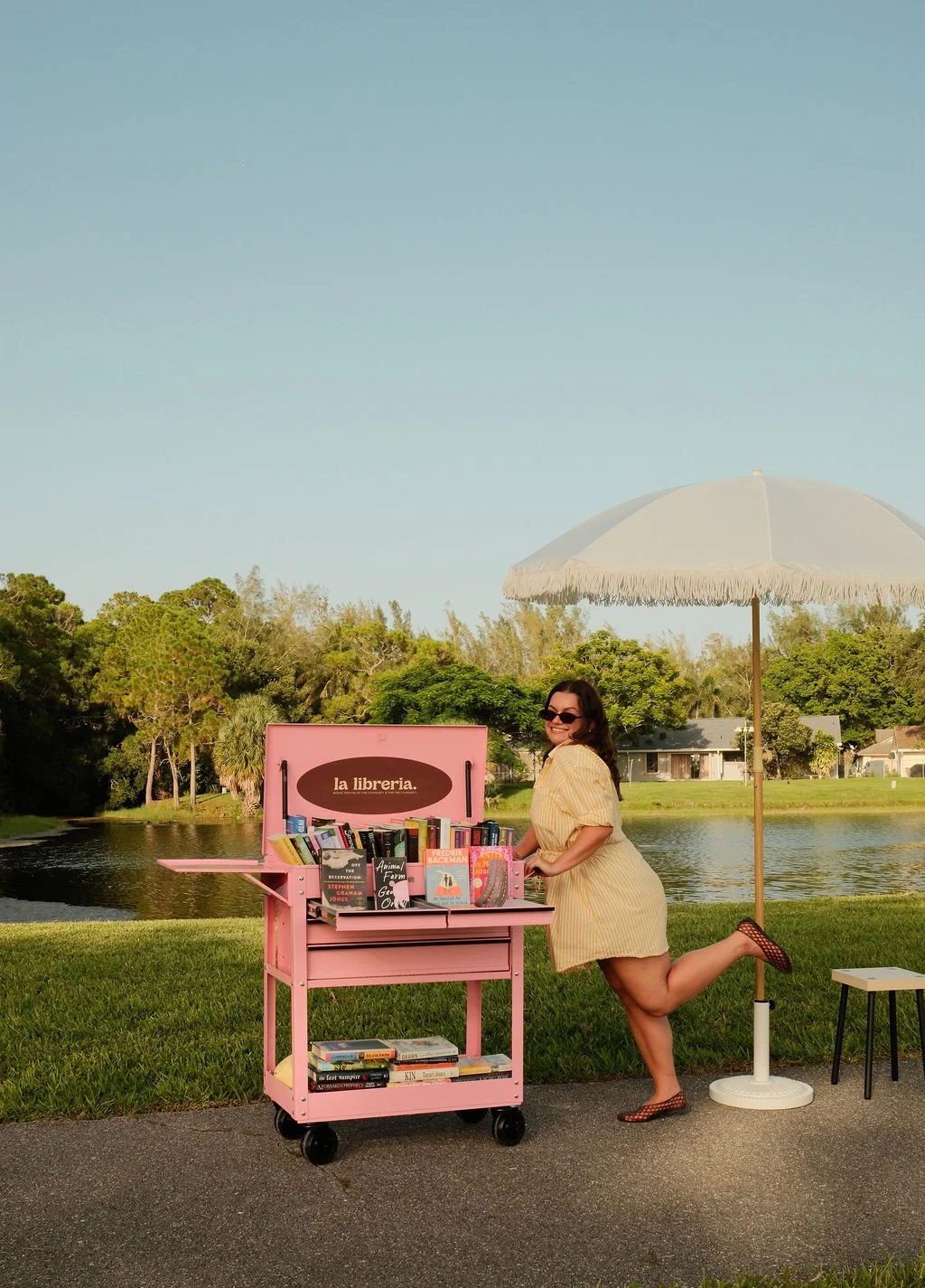 A woman in a yellow dress and sunglasses standing beside a pink mobile library cart with books, by a pond with green trees and houses in the background, next to a large white patio umbrella.