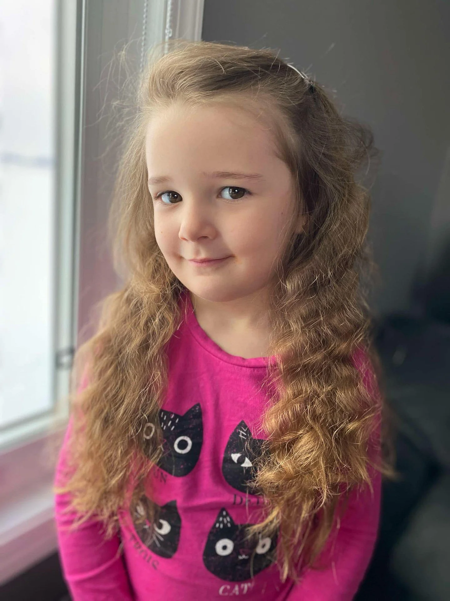Mischievous Grin - Darlene Crawford - LPC / Everest 2024 Competition - Best Portrait. A young girl with long wavy hair and a pink cat-themed shirt smiling gently in soft window light. #prpacanada #prpa #photography #portrait
