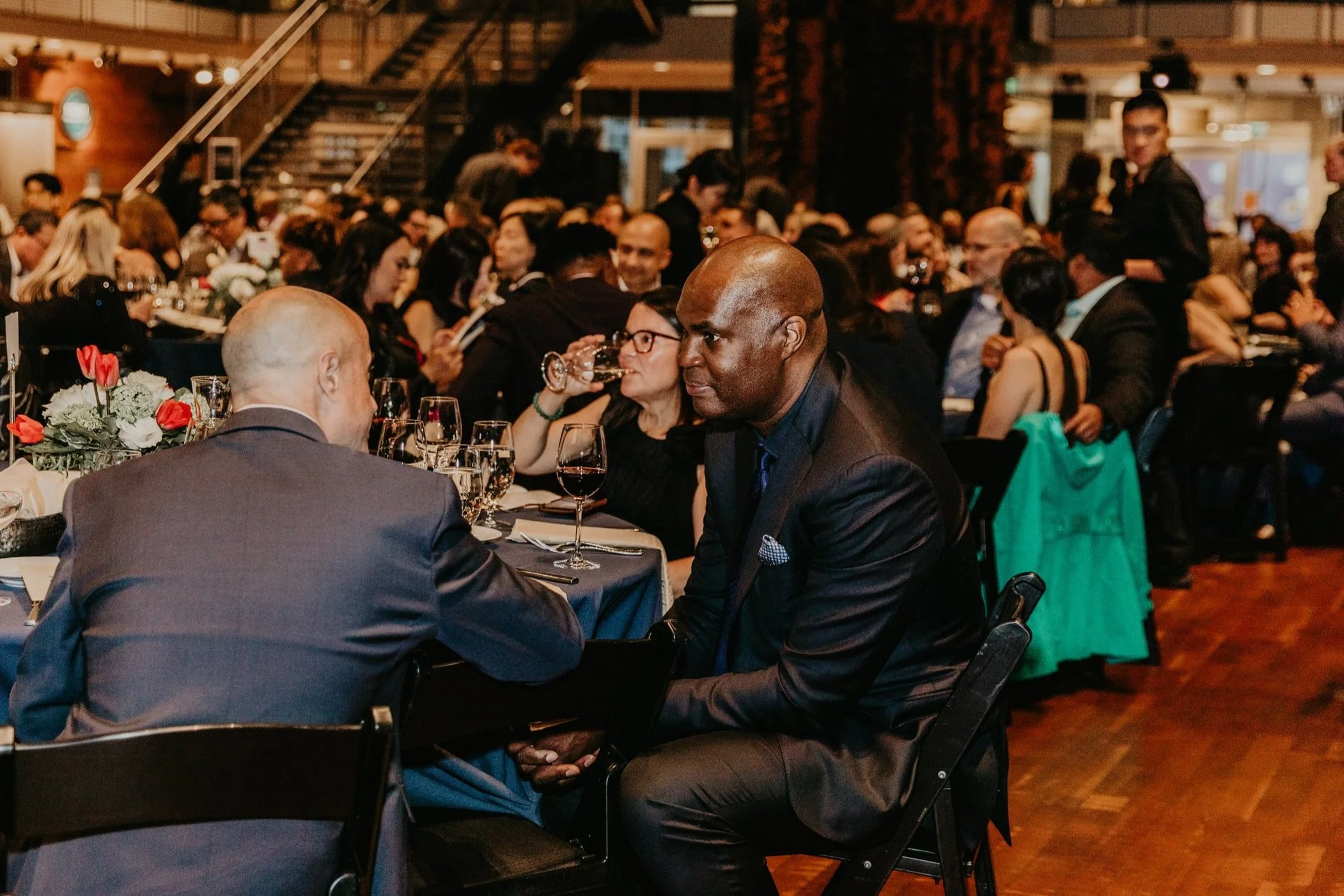 People sitting at a formal event, dressed in suits and dresses, engaged in conversation, with tables set with wine glasses and floral centerpieces.