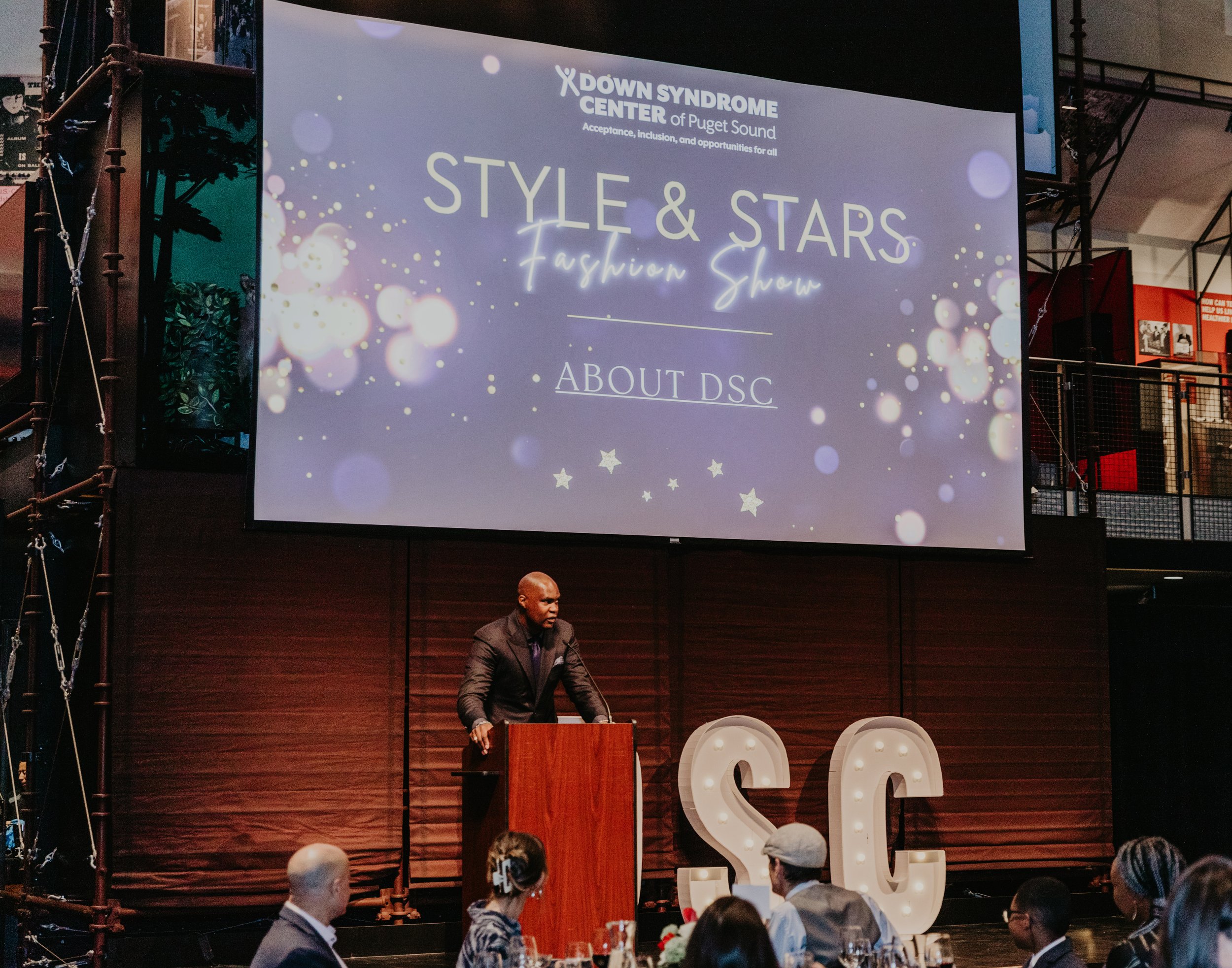 A man speaking at a podium during the "Style & Stars Fashion Show" event, organized by the Down Syndrome Center of Puget Sound. A large screen behind him displays the event's name with decorative lights. "DSC" letters are illuminated on stage, and people are seated at tables.