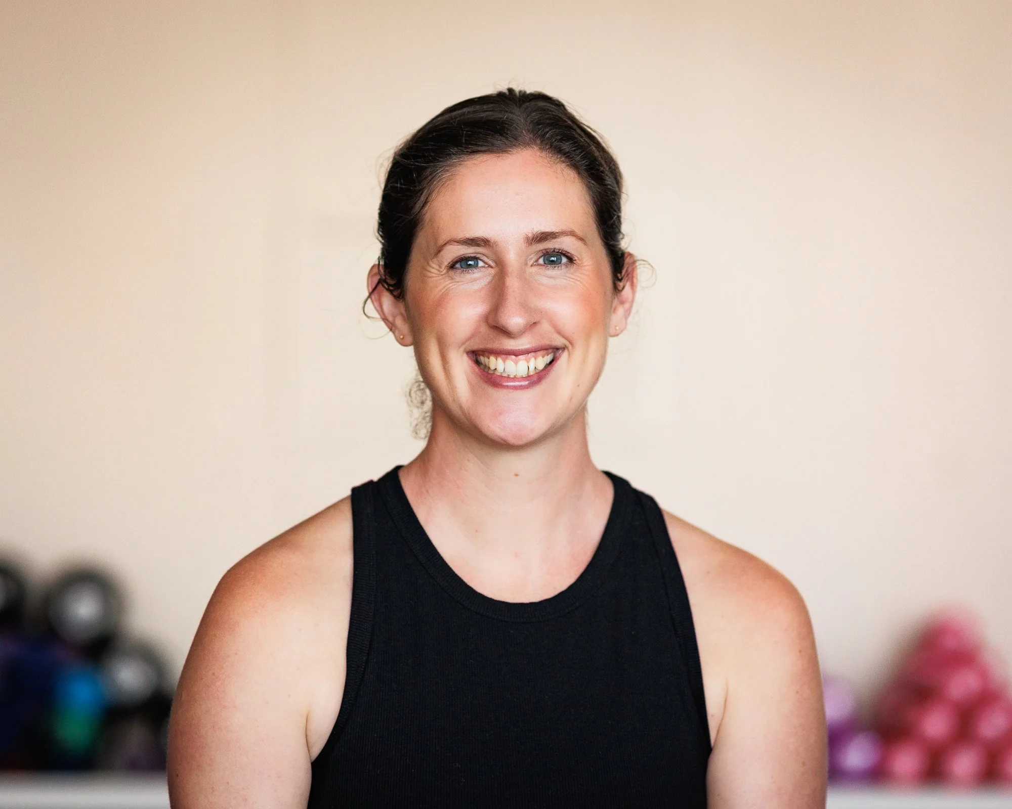 A smiling woman with blue eyes and dark hair tied back, wearing a black sleeveless top, in a gym with exercise weights in the background.