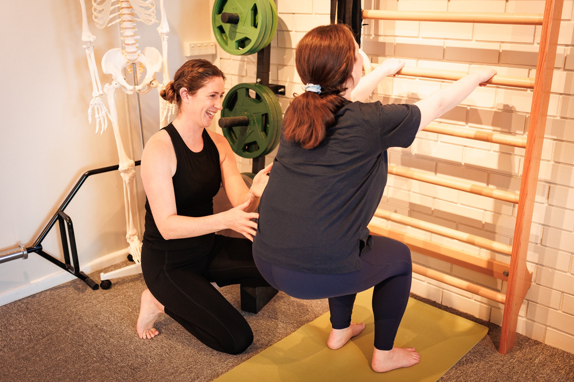 Two women practicing yoga, one assisting the other as she holds a plank pose against a wall-mounted wooden ladder with gym equipment and a skeleton model in the background.