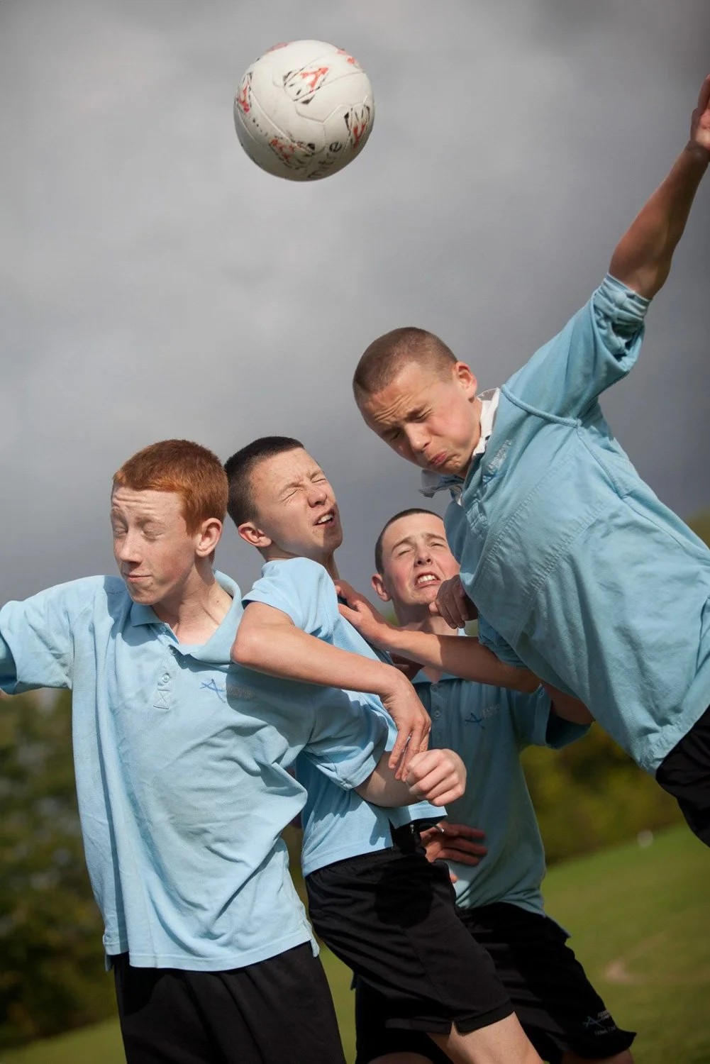 Group of boys in light blue shirts playing soccer, reacting to a ball in the air.