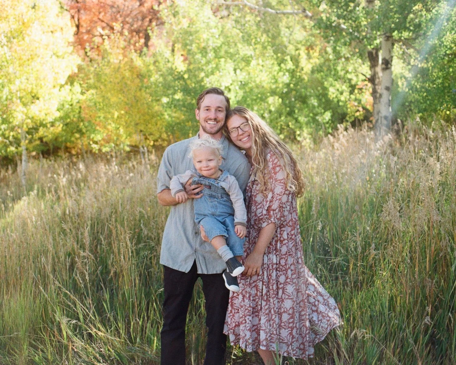 A happy family of three standing in a grassy field with green trees in the background, enjoying the outdoors on a sunny day.