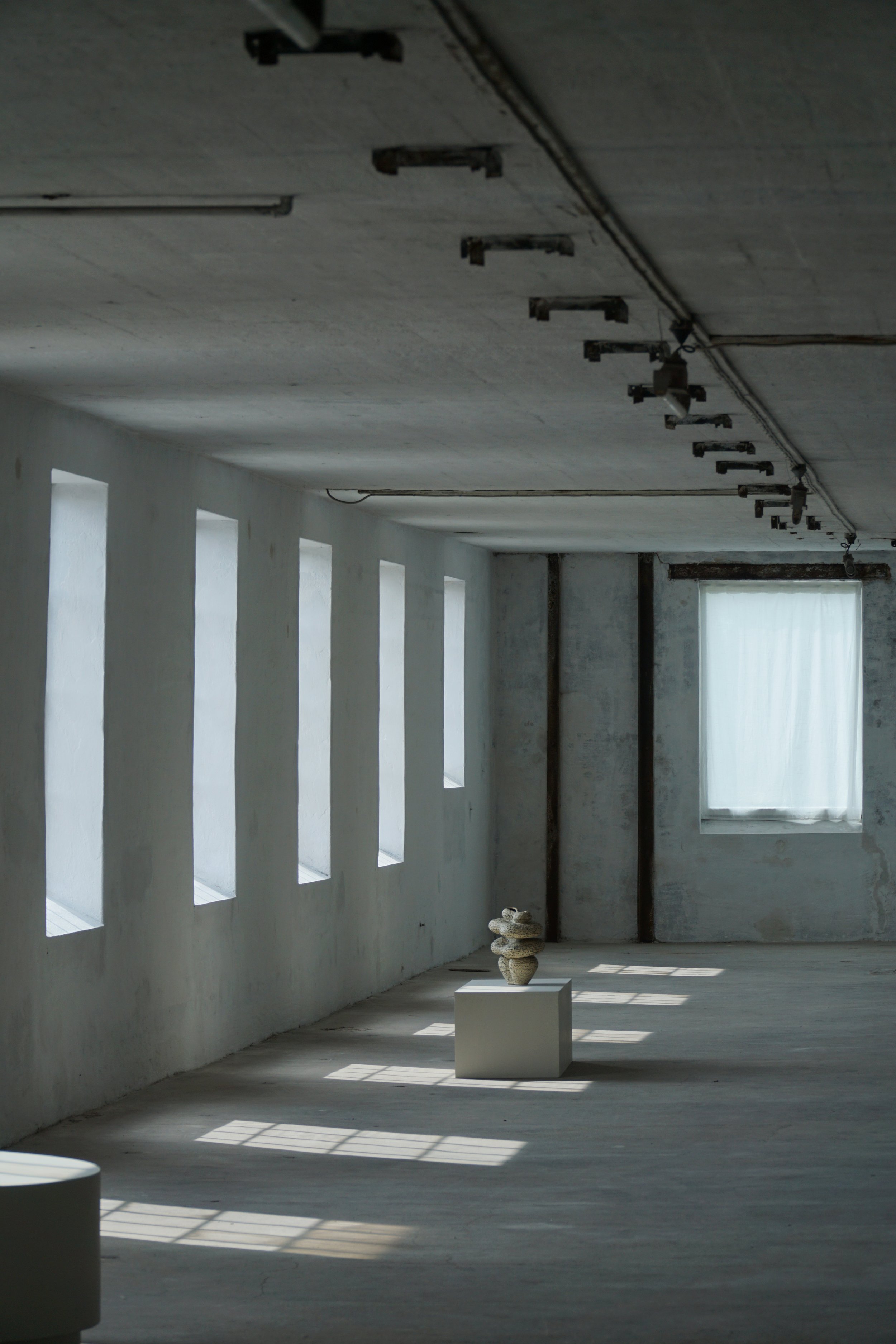 Minimalist art gallery with sunlight streaming through tall narrow windows, a sculpture on a pedestal, and a single large curtain covering a window.