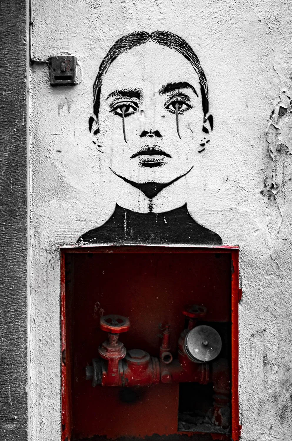 alexander_prior_photographer_vancouver_Graffiti of a woman's face with tears on a textured wall, positioned above a red fire hose cabinet.