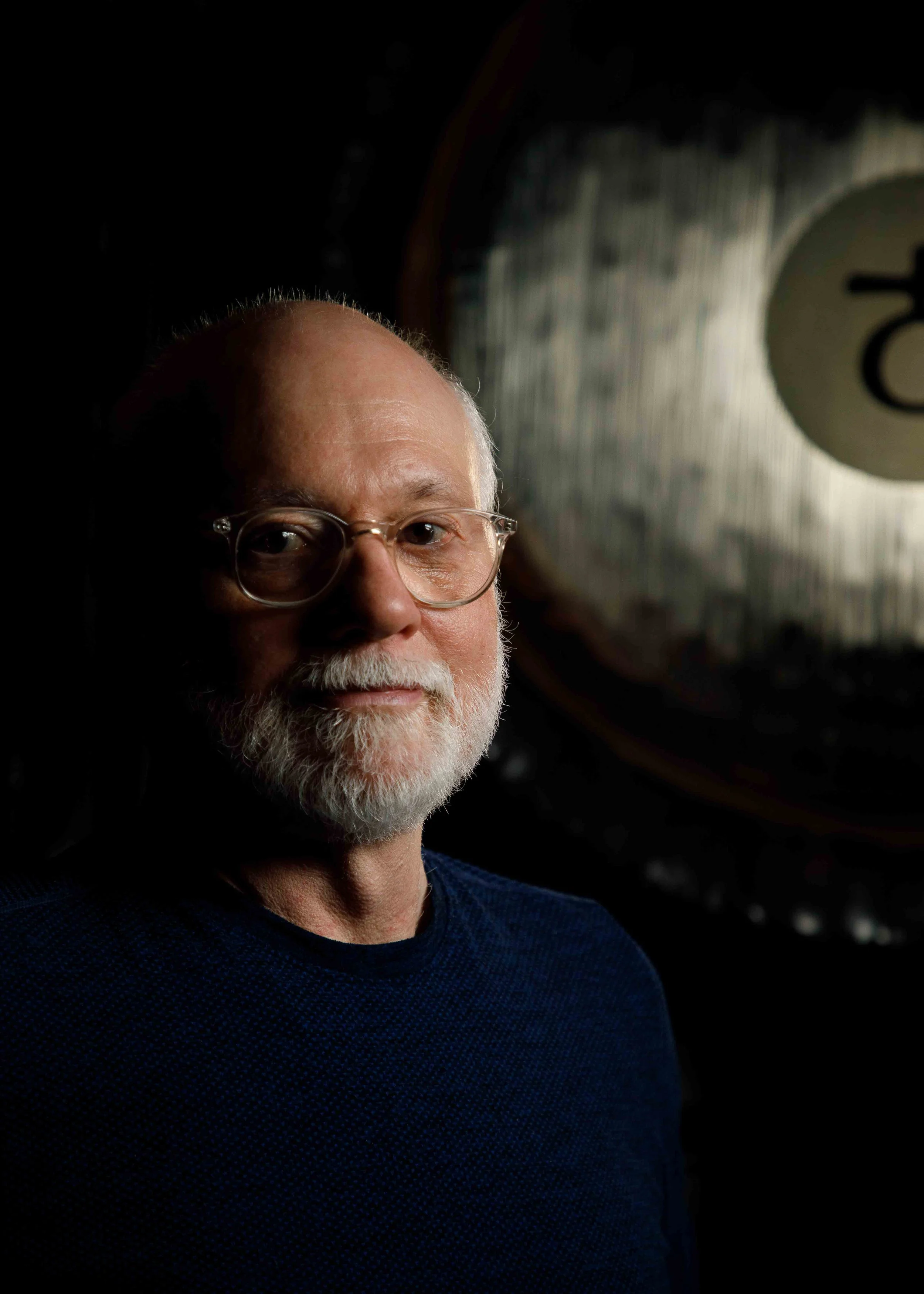 alexander_prior_portrait_photographer_vancouver_portrait of an older man with glasses, a beard, and light complexion, looking at the camera, in front of a dark background with a gong behind him.