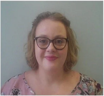 Head-and-shoulders photo of a Health Liaison Team member wearing glasses and a patterned top, photographed against a plain indoor wall.