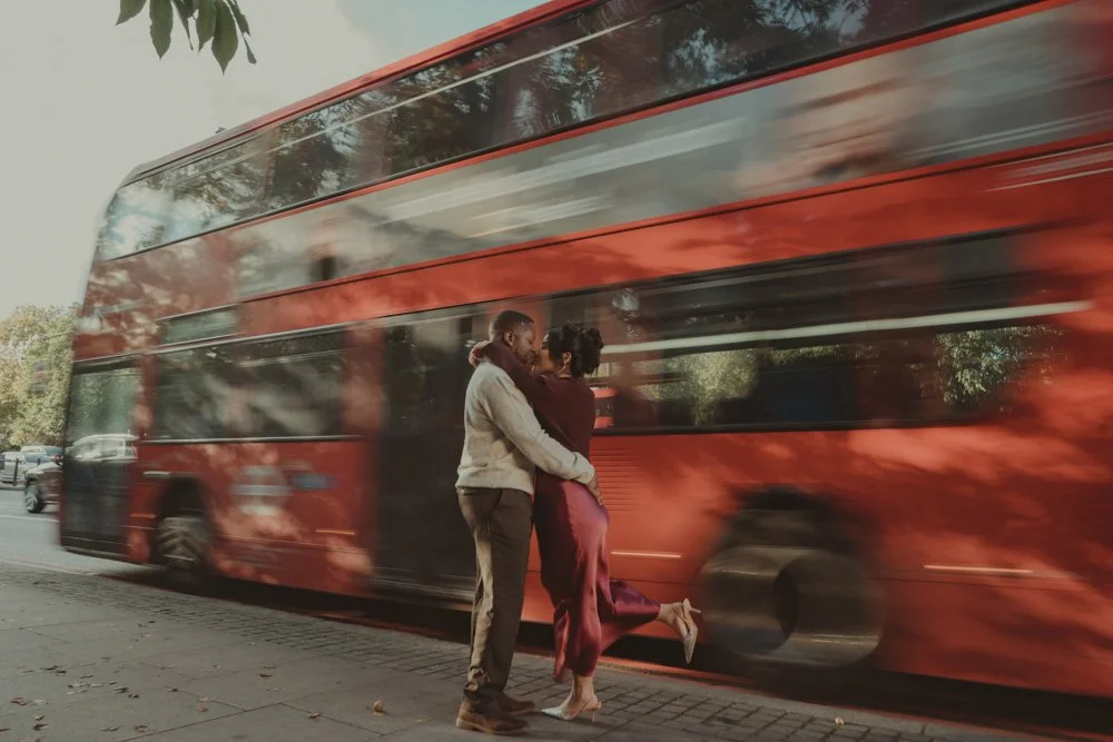 Long exposure photo of a couple kissing, with a red bus passing behind them, on the streets of London
