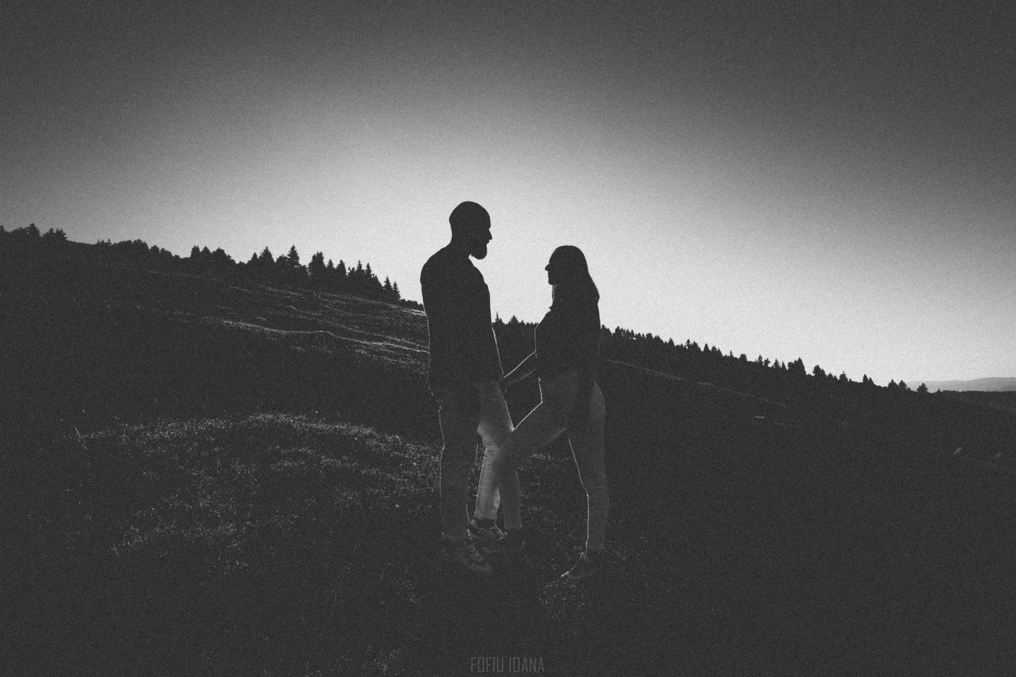 Black and white contre jour photo of a couple looking at each other while holding hands on a hill 