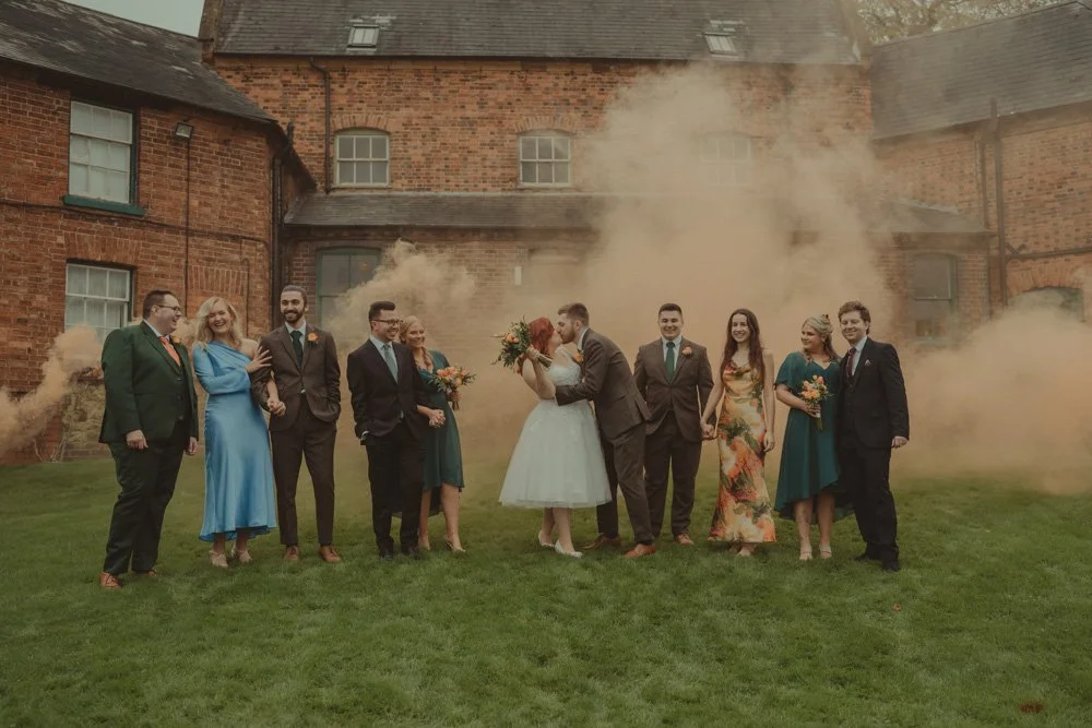 Bride and groom kissing, surrounded by friends at their wedding, for a smoke bomb group photo, at The Barns at Hunsbury Hill