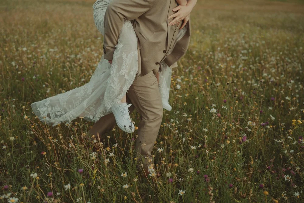 Groom walking through flowers on a field, carrying the bride on his back, at Wellington Barn, Wiltshire