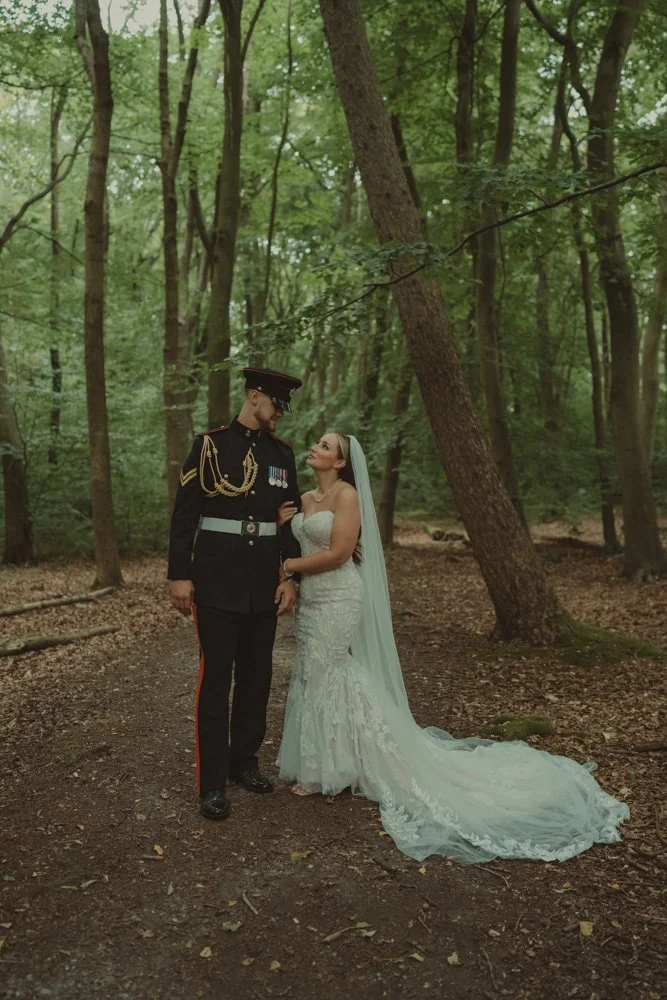 Groom in military costume facing the camera, and bride holding his arm and looking at him, in the woods of Basingstoke, UK
