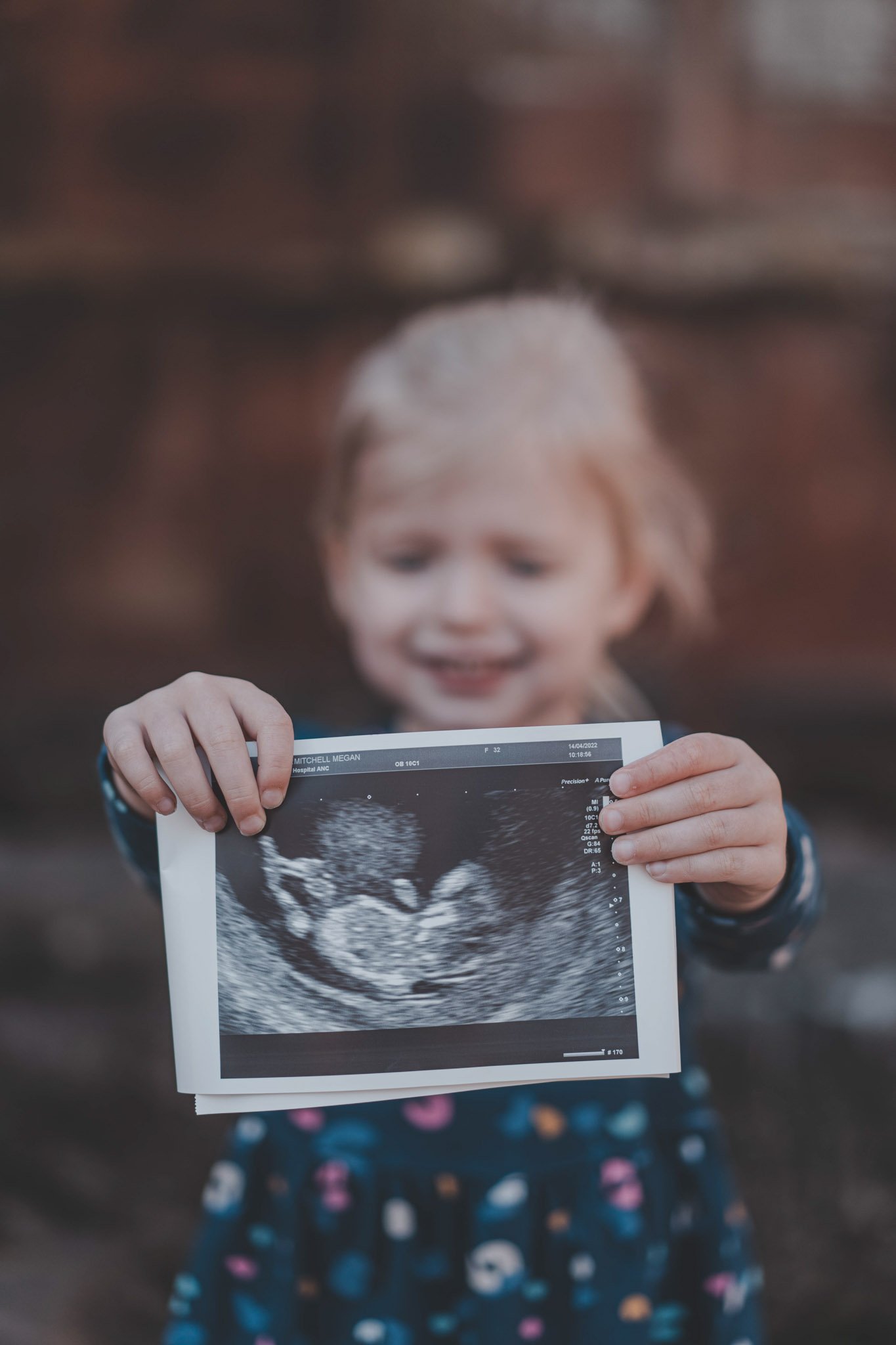 Small girl smiling while photographed holding a  ultrasound scan photo of her future brother