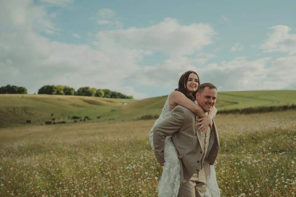 Groom carrying the bride on his back, laughing together, on the fields of Wellington Barn, Wiltshire