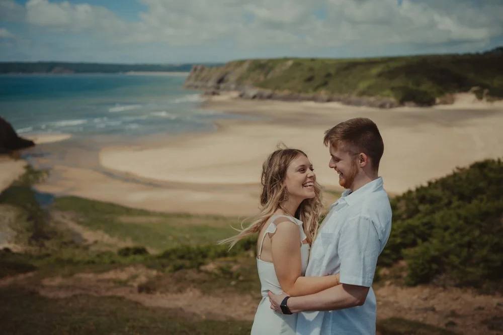 Couple tickling each other and laughing during a save the date photo session, with Three Cliffs Bay in the background 