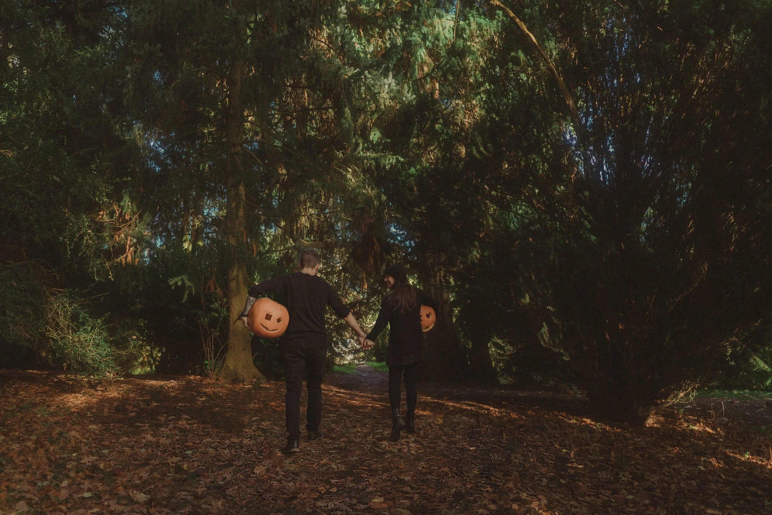 Couple wearing black, holding two carved pumpkin heads, walking away from the camera holding hands, at Coombe Abbey Park in Coventry