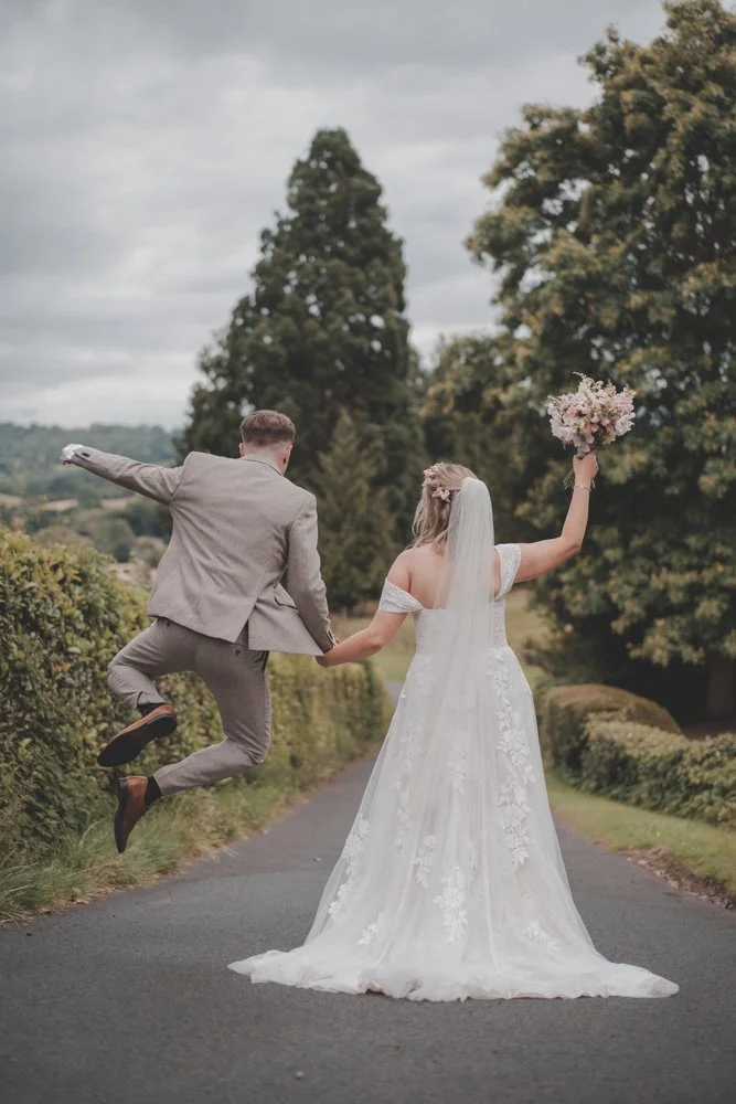 Funny photo of a bride and groom walking with their backs to the camera, groom jumping in the air, at The Courtyard at Shelsley Walsh