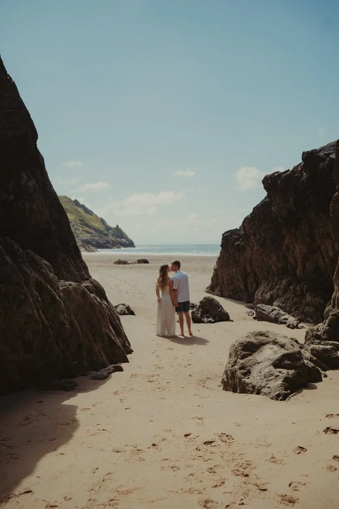 Vertical photo of a couple waring white, with their back towards the camera, at Three Cliffs Bay, Wales