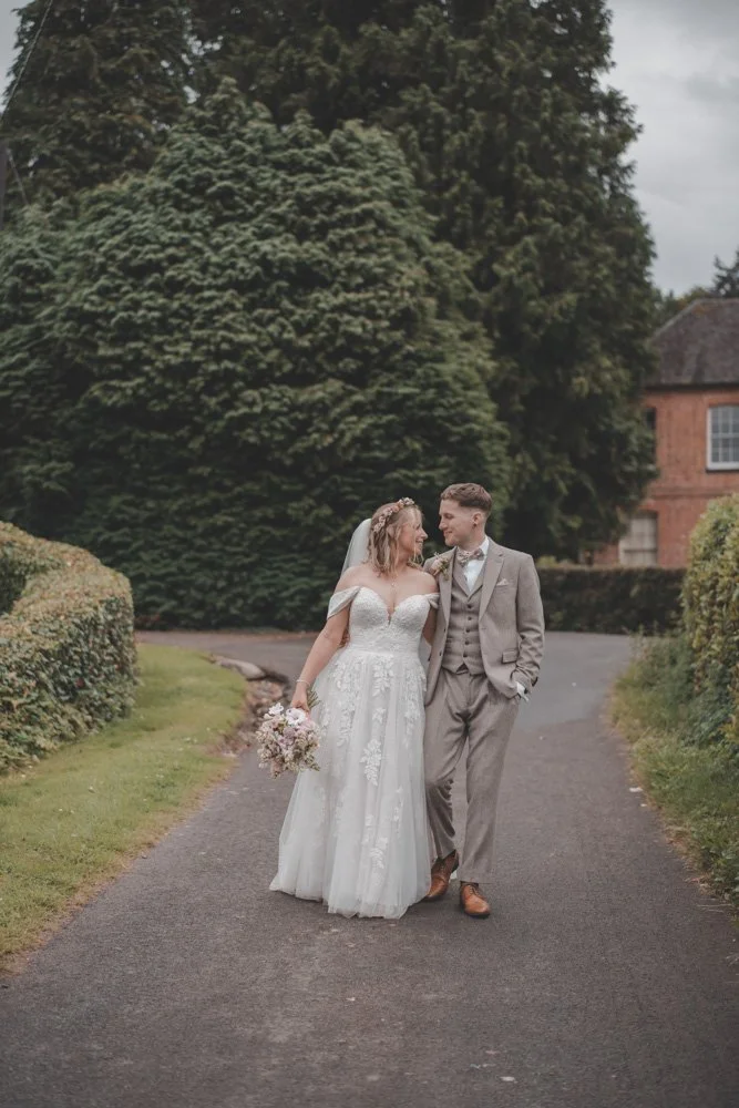 Vertical full body portrait of bride and groom holding and looking at each other, walking towards the camera on the grounds of The Courtyard at Shelsley Walsh