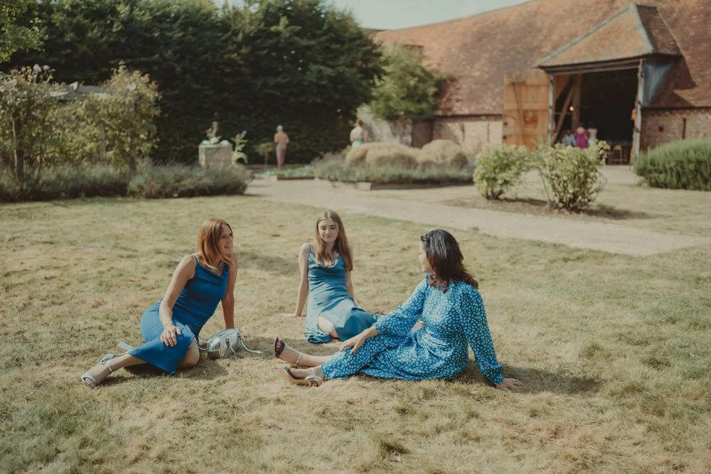 Three women wearing blue, photographed sitting on the grass and having a chat during a wedding, in front of a barn in Drayton, Oxfordshire