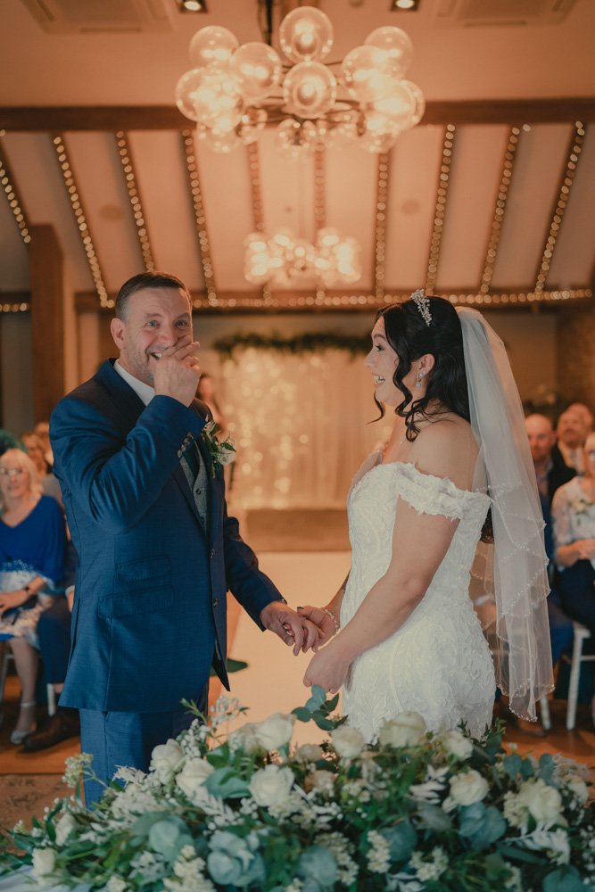 Vertical photo of groom and bride facing each other during their wedding ceremony, with groom crying, at The Waterfront Barton Marina