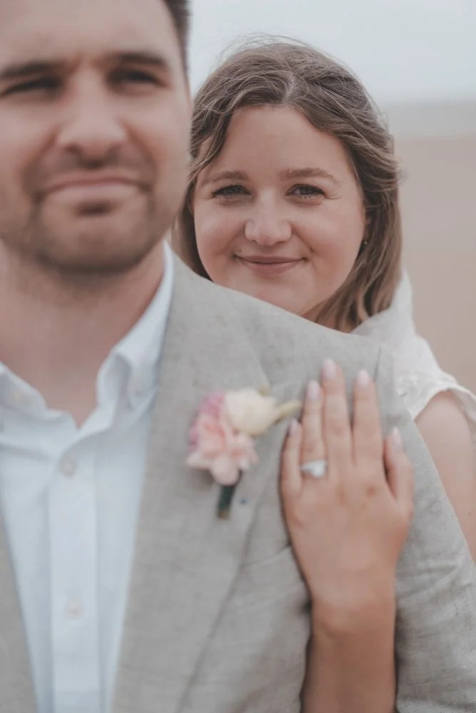 Bride hugging groom from behind, you can see the engagement ring on her finger
