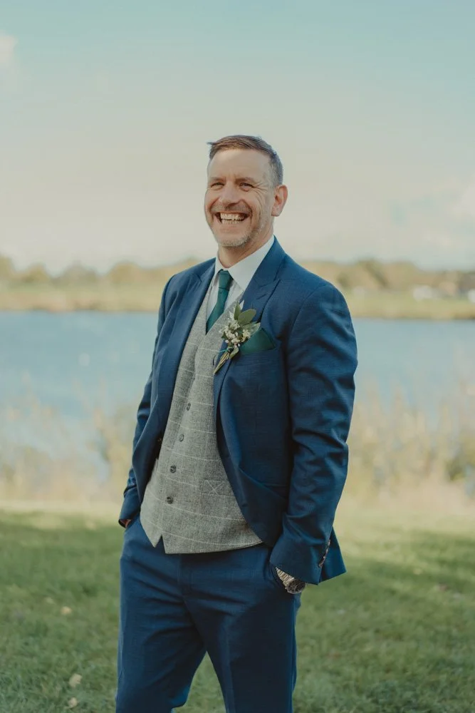 Groom photographed with hands in pocket and laughing, on a sunny wedding day, at The Waterfront Barton Marina