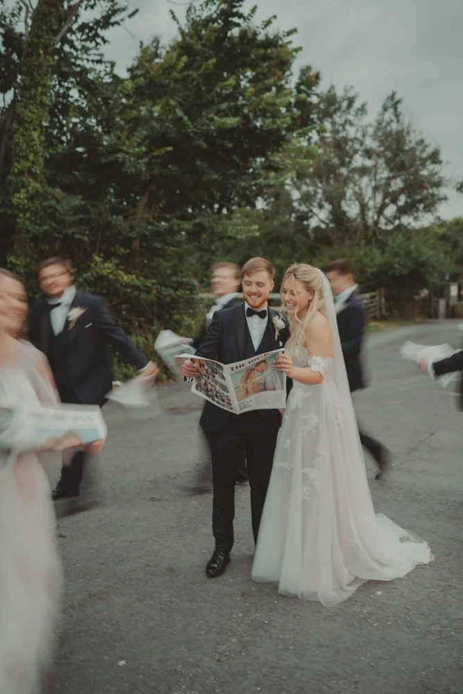 Long exposure photo of bride and groom holding a wedding newspaper, with wedding guests running around them, at Gwesty'r Emlyn Hotel in Newcastle, Wales