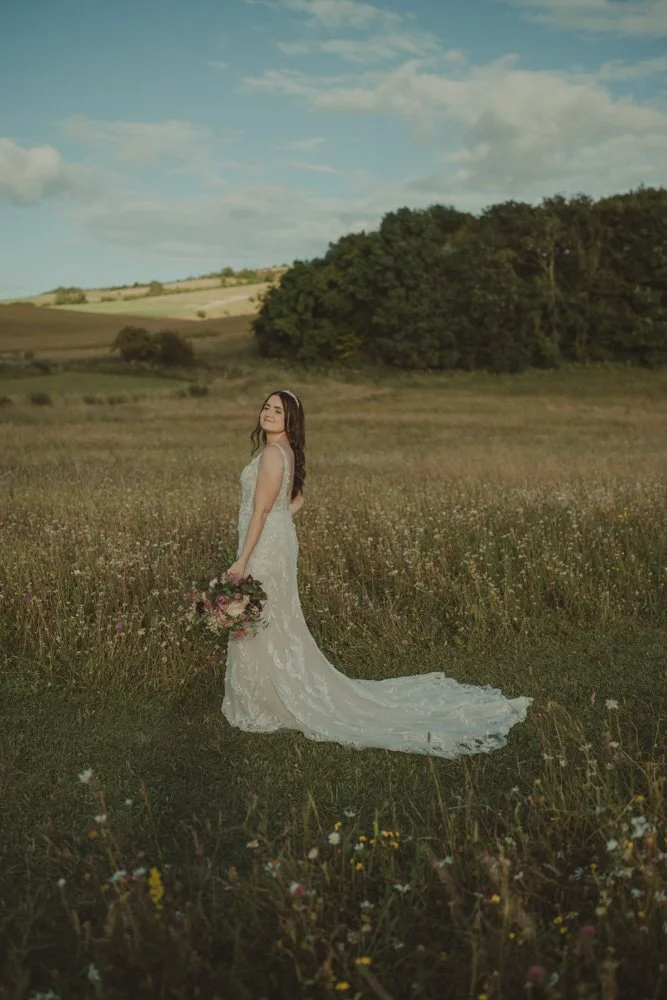 Vertical photo of a bride holding the flowers in a hand and looking at the camera over the shoulder, on the grounds of Wellington Barn, Wiltshire