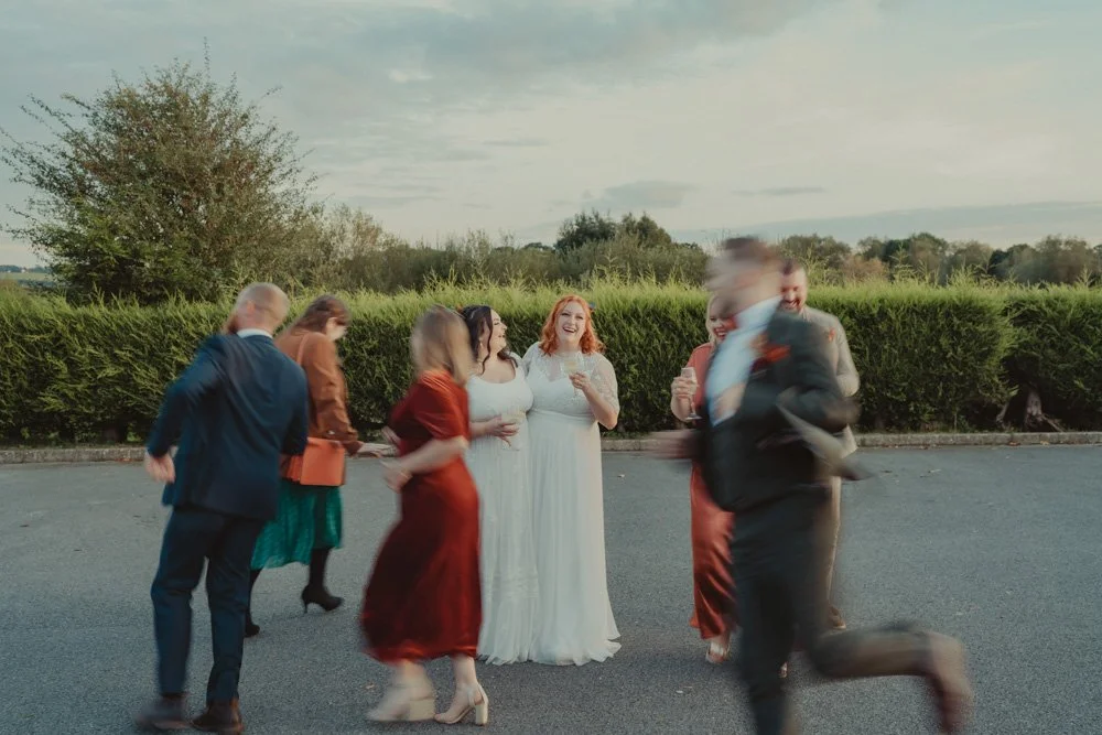 Photo of two brides laughing, while friends run in a circle around them, for a long exposure photo, at The Chester Fields Country Pub, Restaurant & Wedding Venue