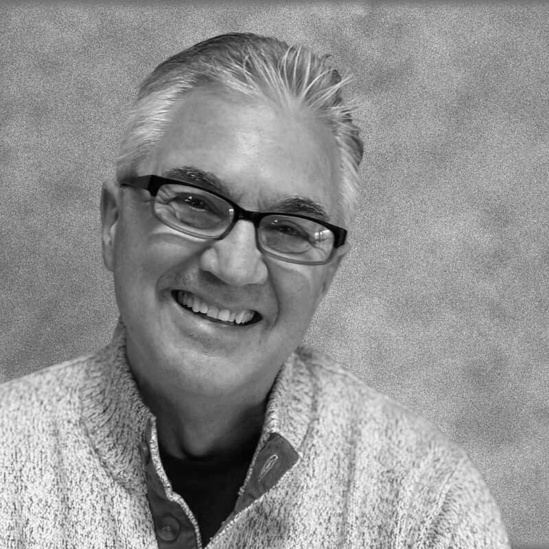 A smiling man with glasses and gray hair, wearing a textured sweater.