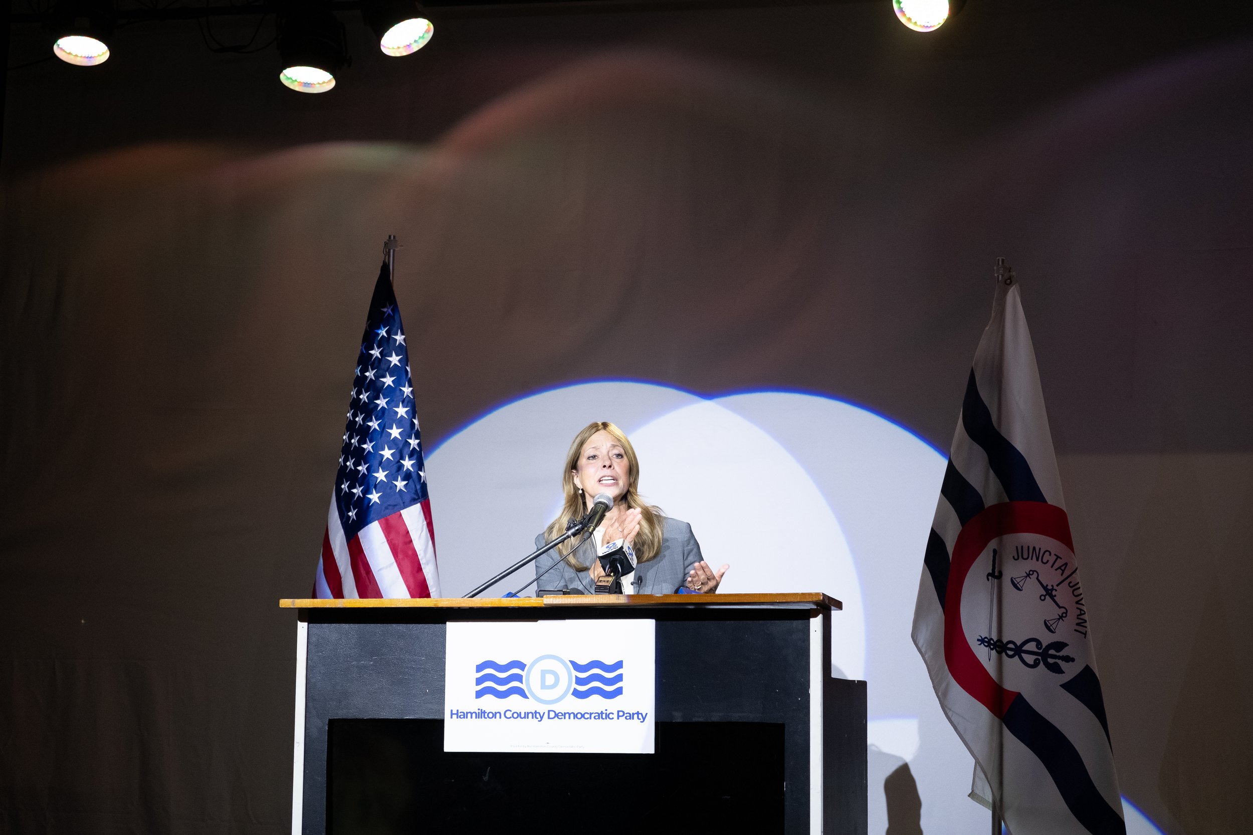 A woman speaking at a podium with signs indicating Hamilton County Democratic Party, flanked by flags of the United States and Hamilton County. The backdrop is dark with overhead lights.