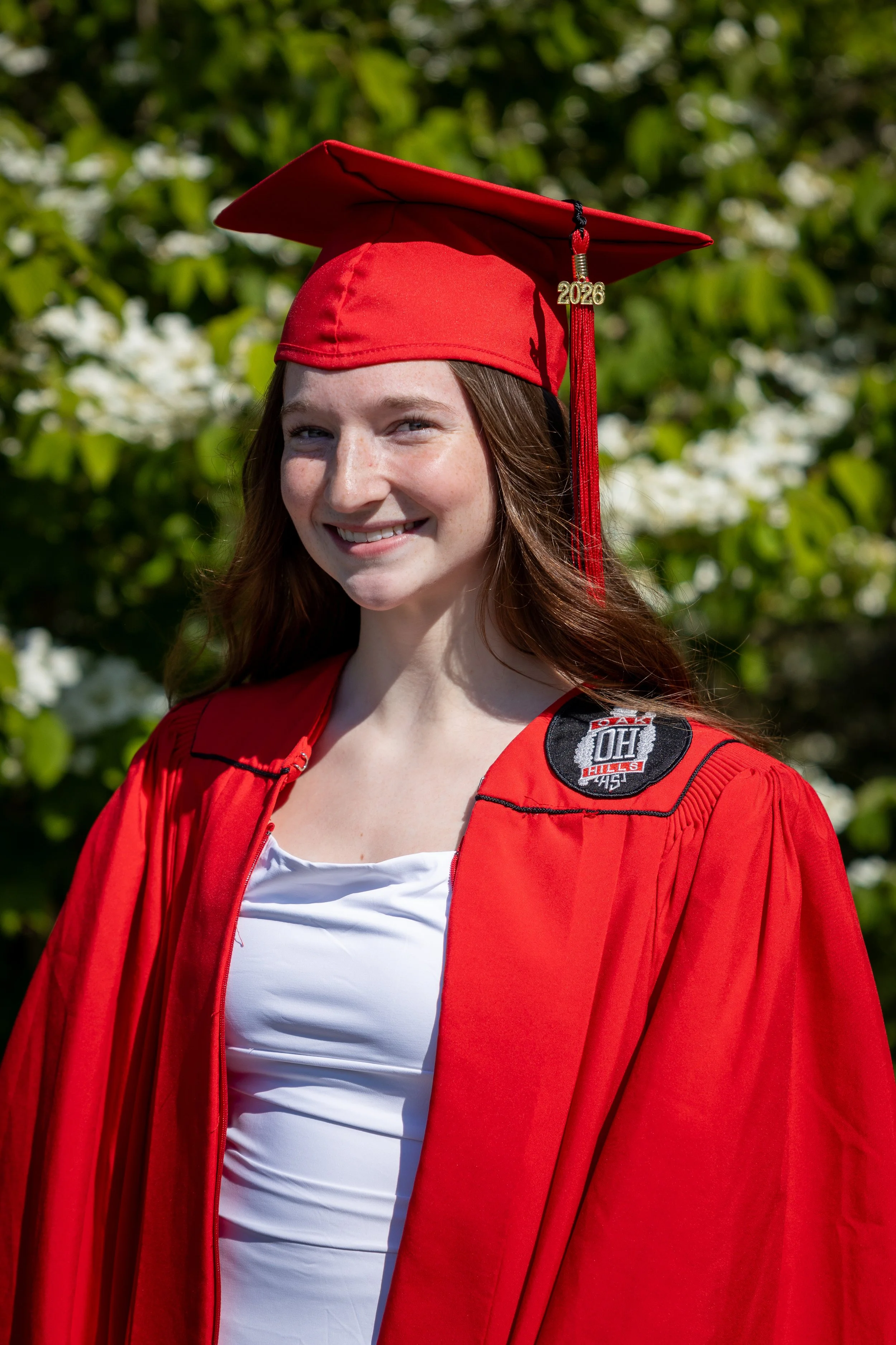 A young woman wearing a red graduation cap and gown with a year pendant hanging from the cap audience in a garden setting