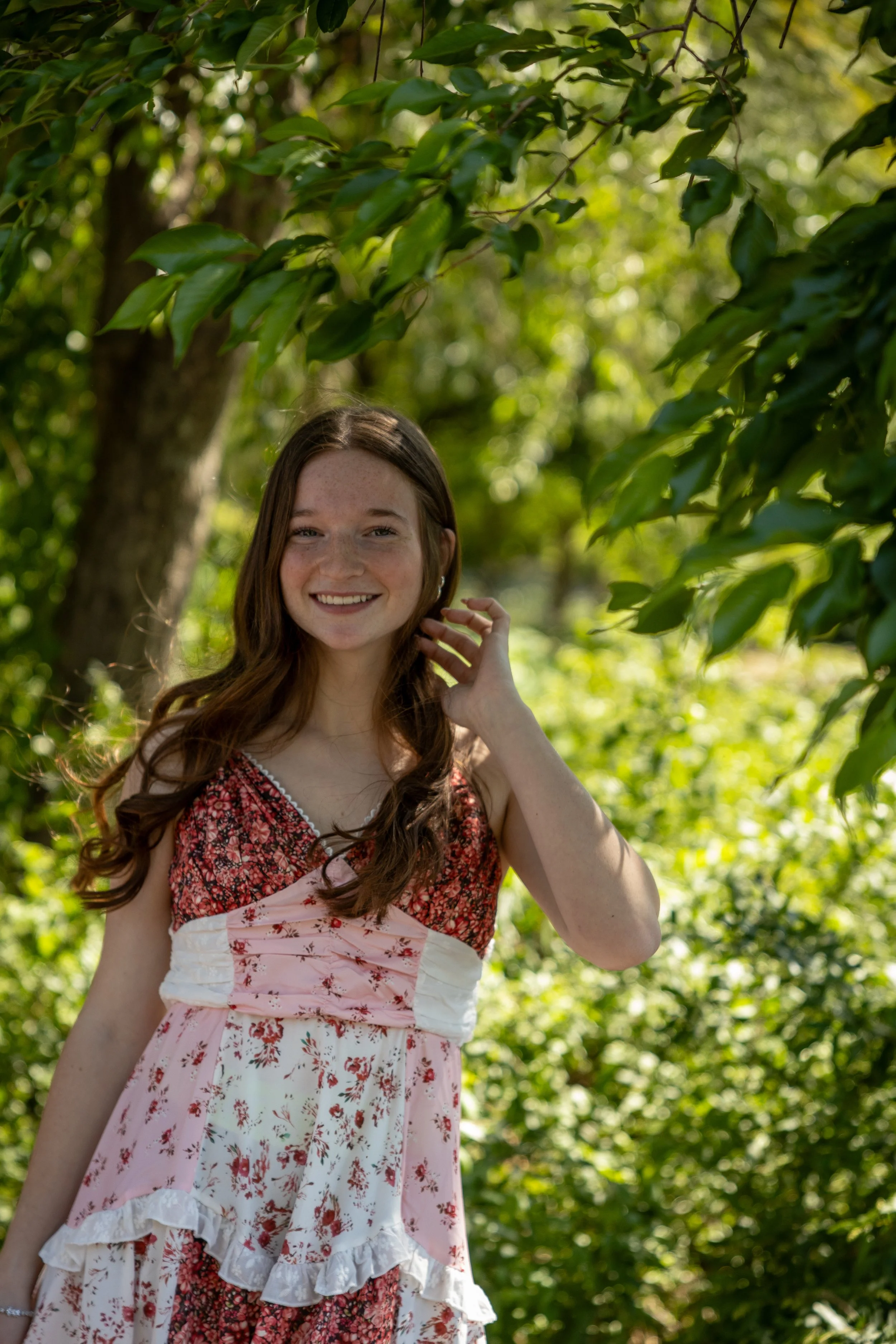 A young woman with long wavy brown hair and freckles, smiling and touching her hair, outdoors among green trees and sunlight filtering through the leaves.