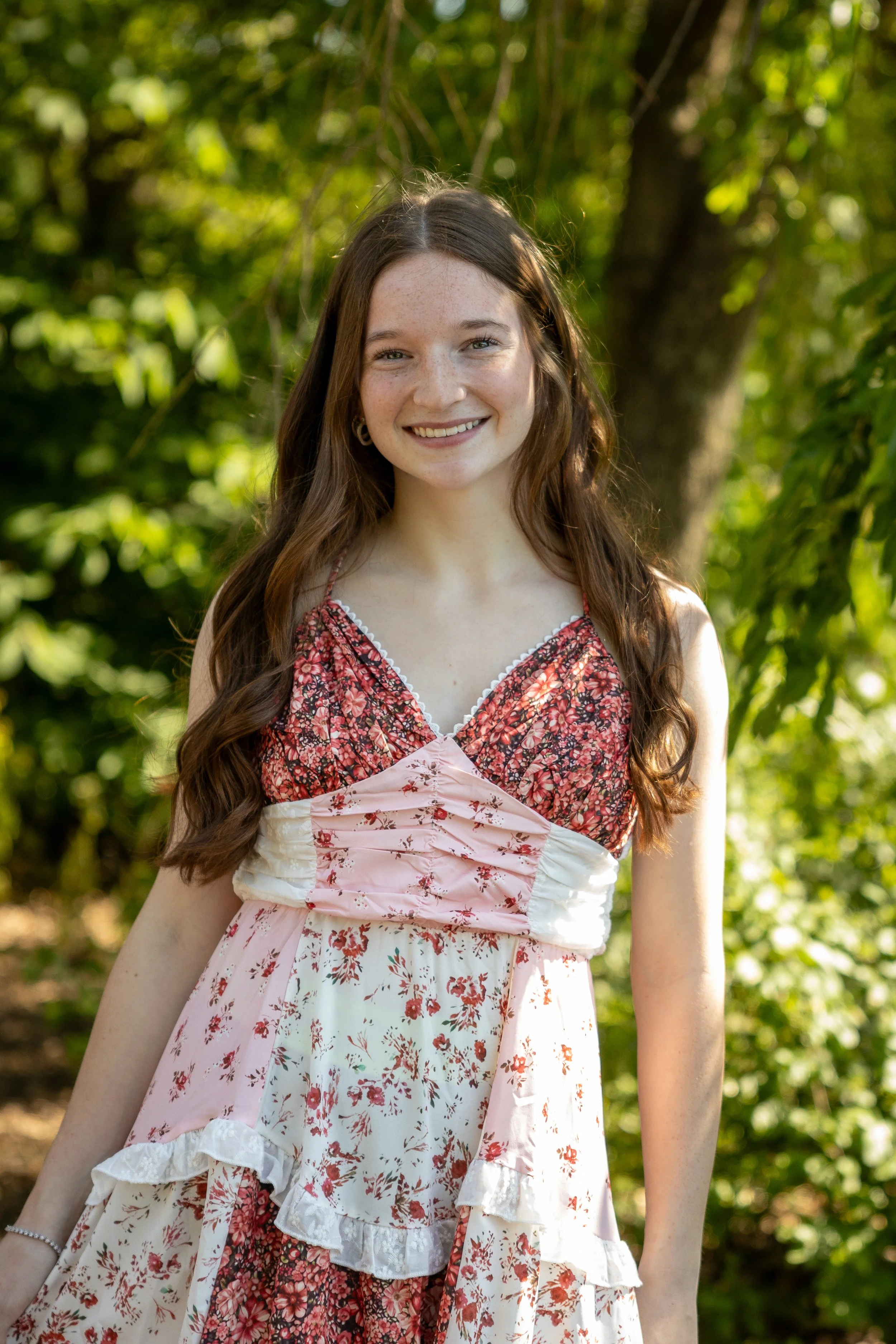 A young woman with long brown hair, wearing a floral dress, is standing outdoors with a background of green trees and foliage, smiling at the camera.